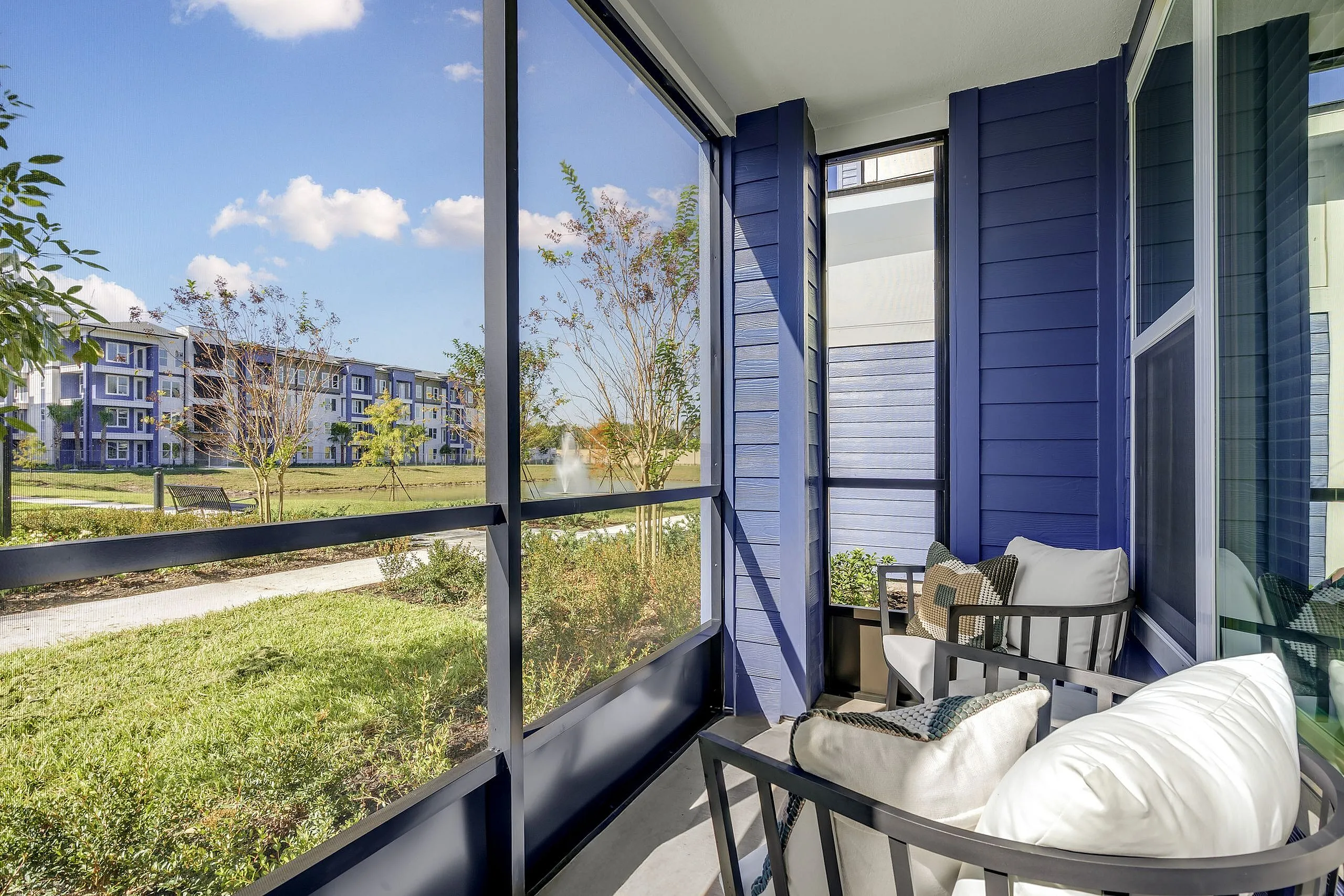 Charming screened balcony with plush seating, overlooking lush greenery and a picturesque pond. Modern blue siding adds a touch of elegance.