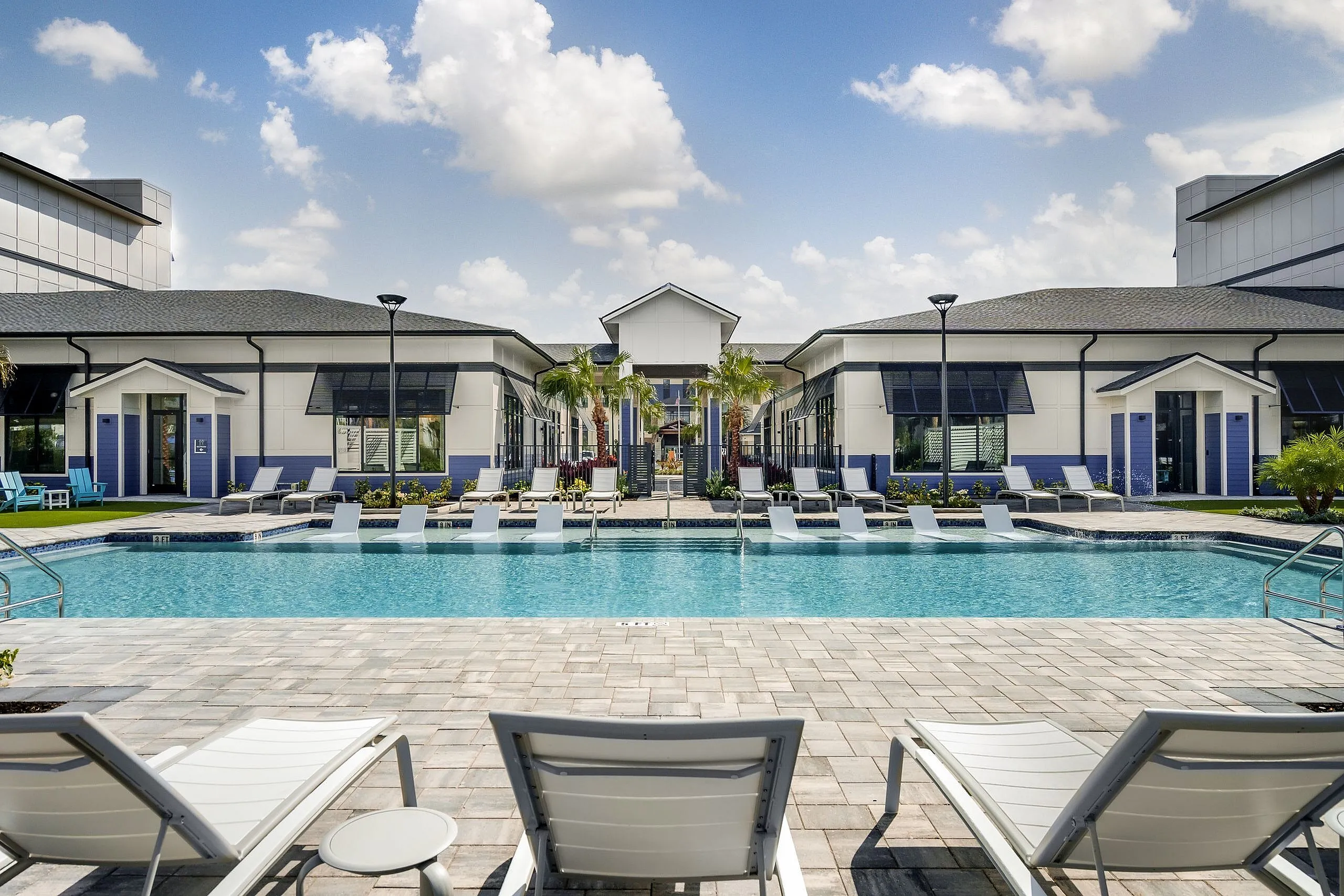 Luxurious apartment complex featuring a stunning outdoor pool surrounded by elegant lounge chairs and modern architecture under a bright blue sky.