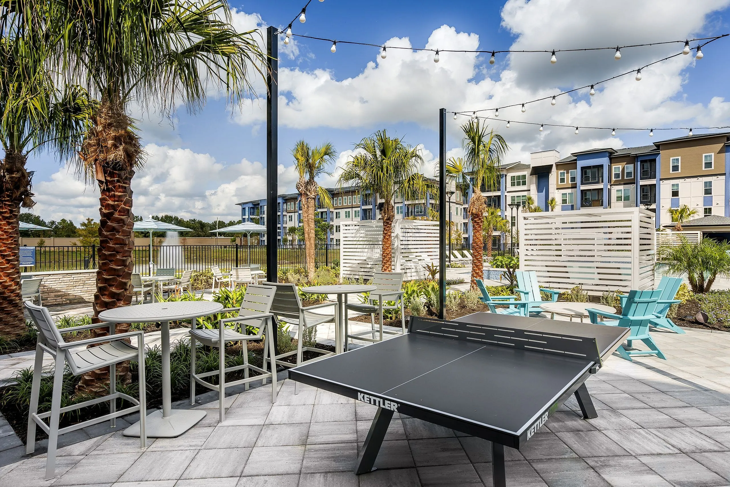 Vibrant outdoor recreation area featuring a modern ping-pong table, comfortable seating, and charming string lights amidst lush palm trees and scenic apartment views.
