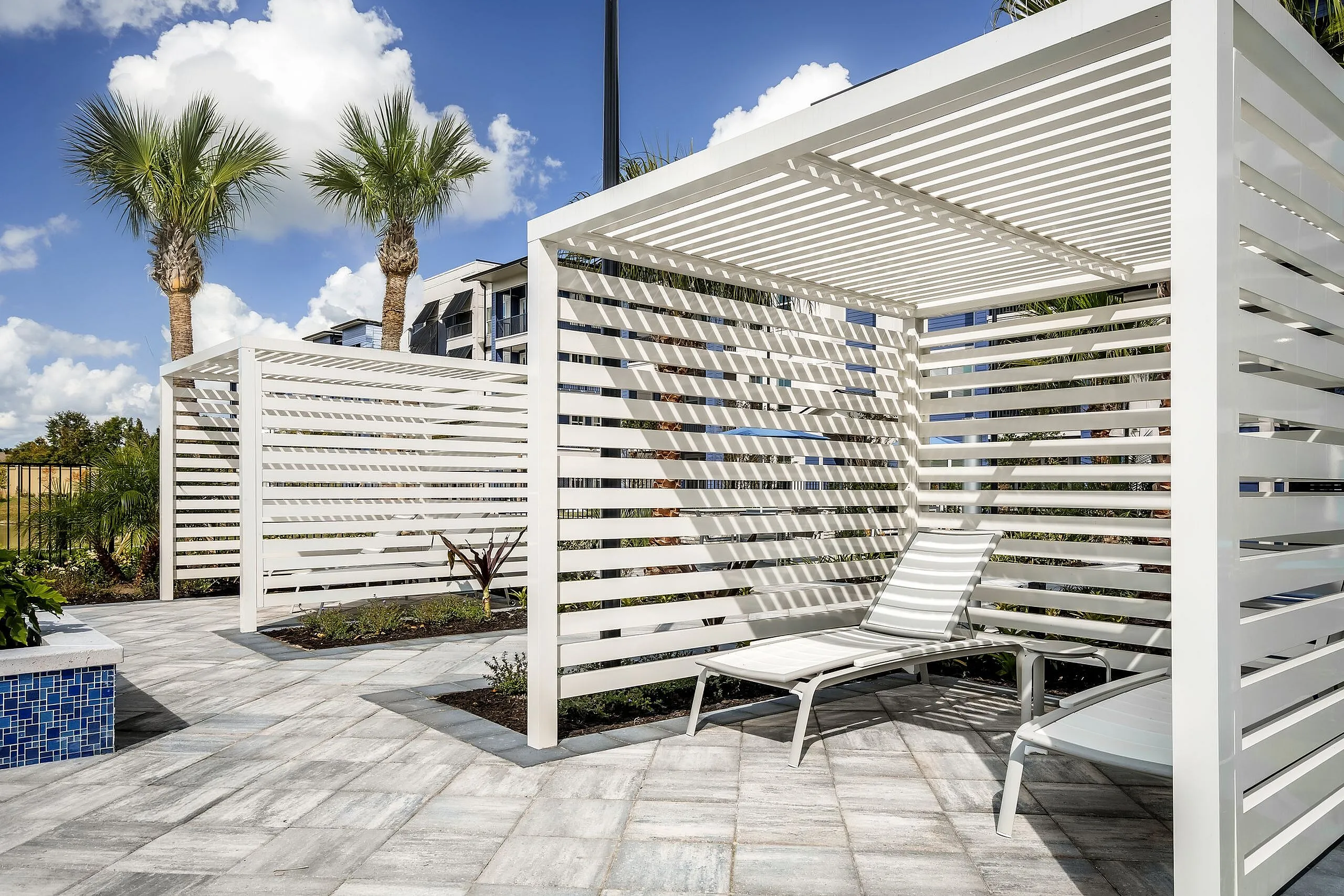 Modern outdoor oasis featuring sleek white pergolas, elegant lounge chairs, and lush palm trees under a bright blue sky.