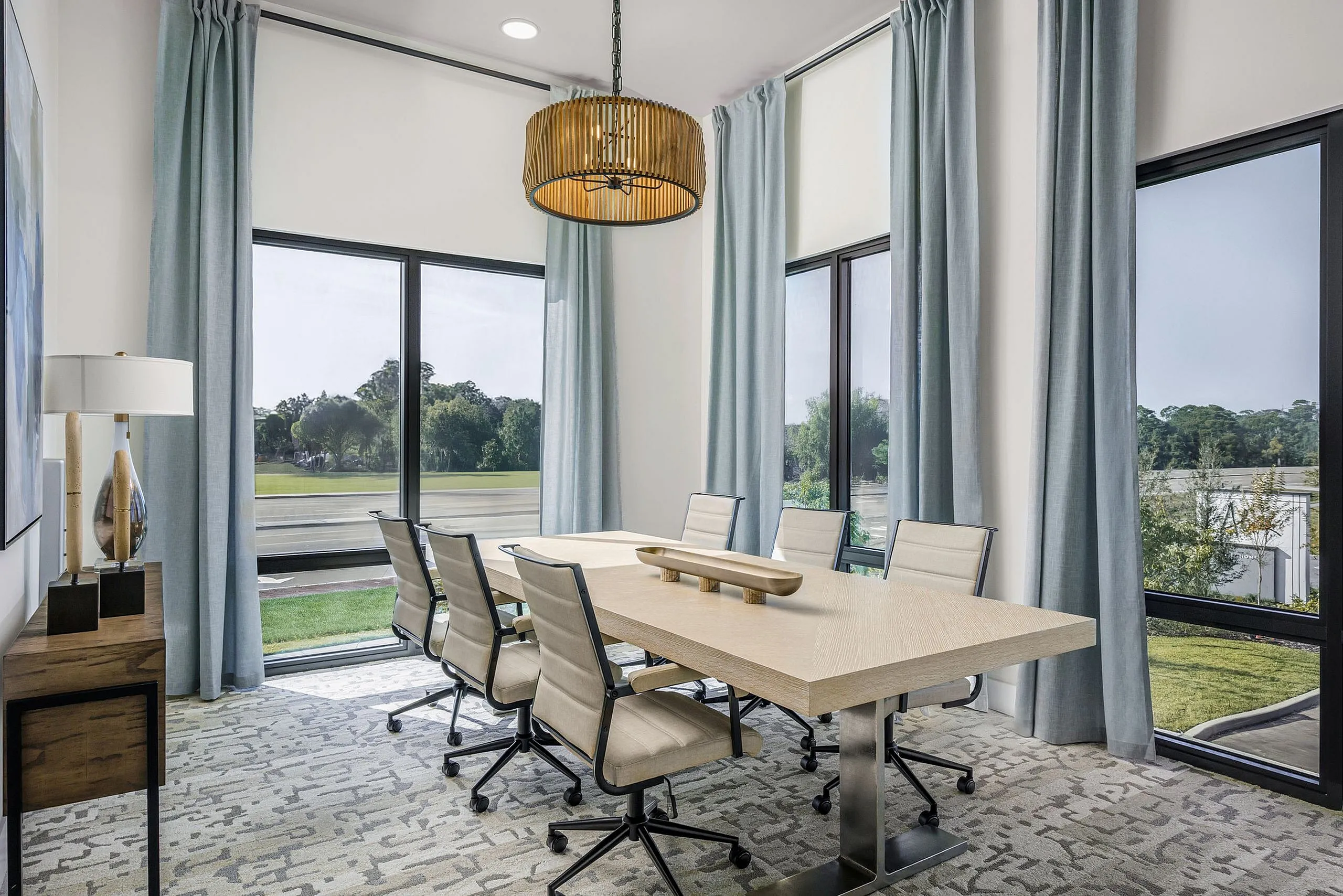 Elegant dining room with large windows, modern wooden table, and soft blue curtains offering a serene view of greenery.