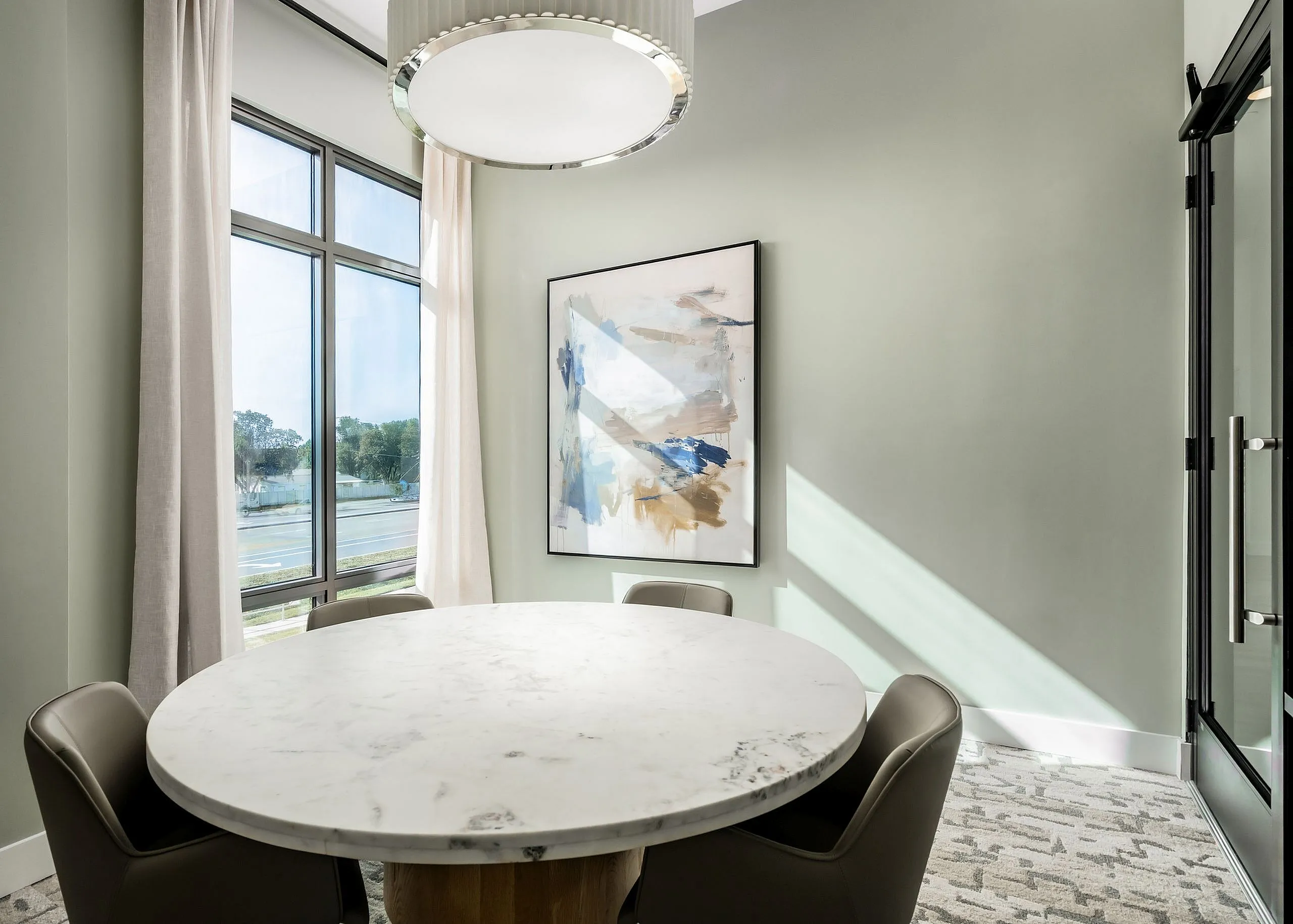 Elegant dining corner with a round marble table, modern artwork, and large windows flooding the area with natural light.