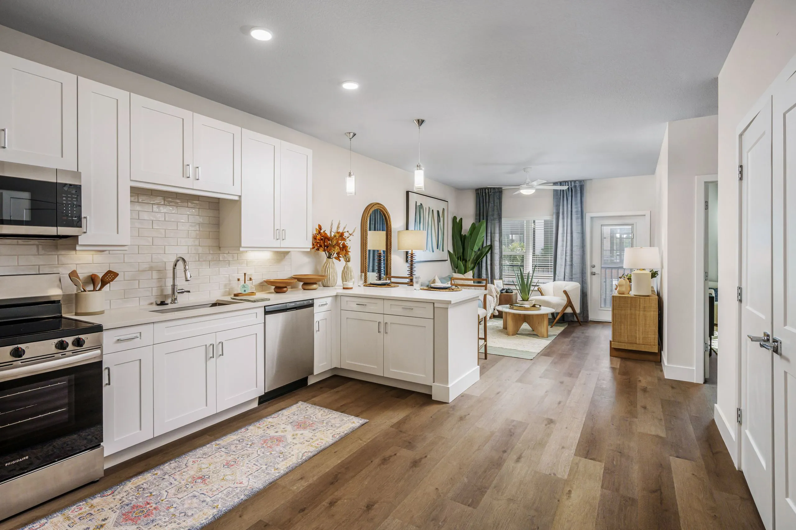Modern kitchen and living space with sleek white cabinets, stainless steel appliances, and warm wooden floors, leading to a bright living room with stylish decor and large windows.