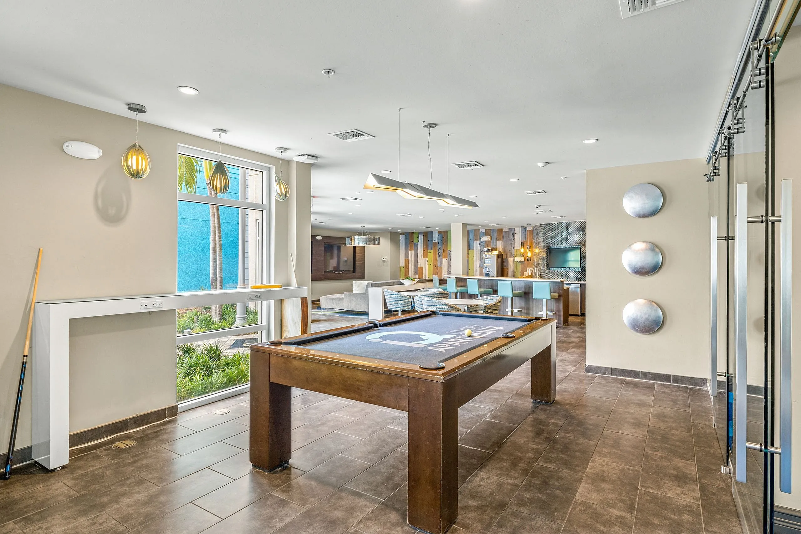 Luxurious apartment lounge with contemporary decor, featuring a pool table, modern lighting, and spacious seating area with a view of palm trees through large windows.