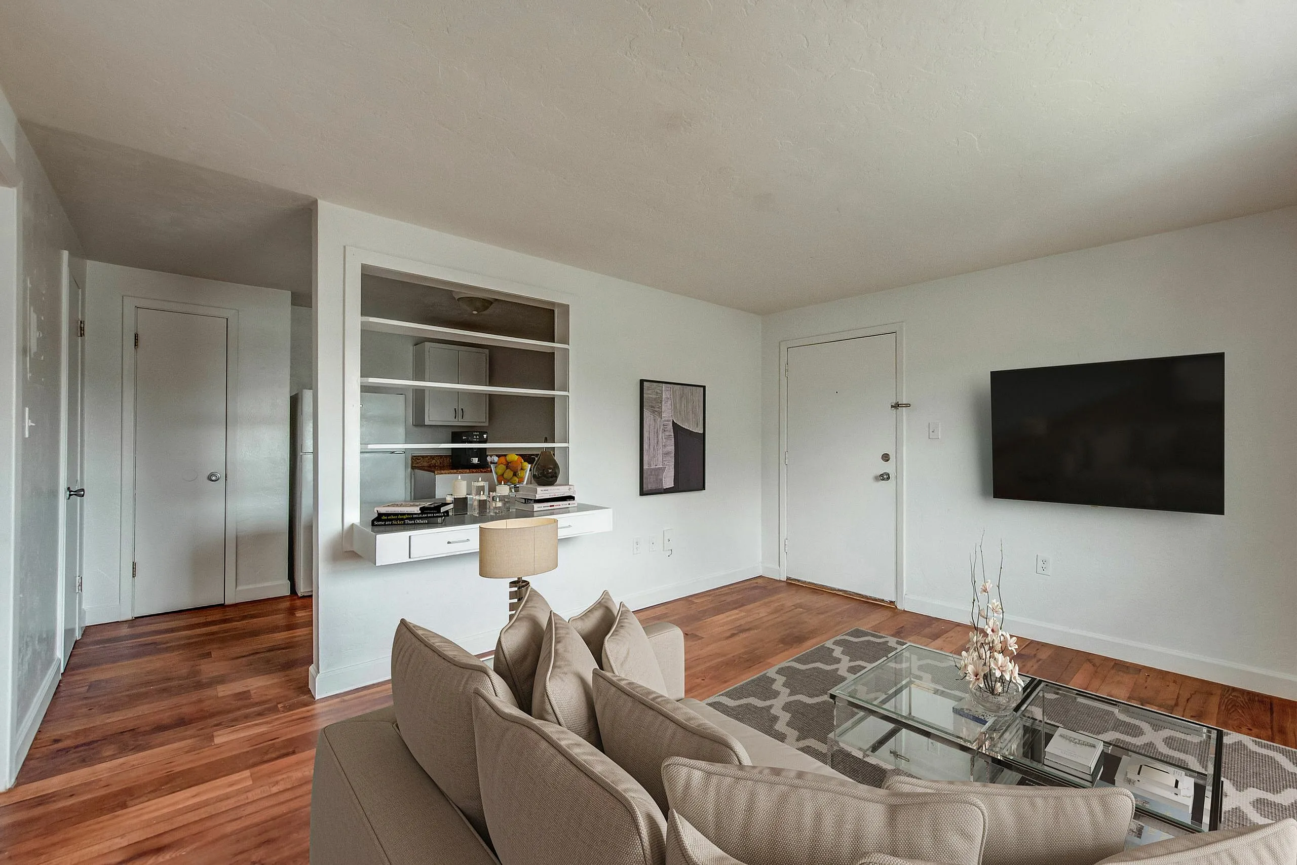 Inviting living room with elegant decor, featuring a sleek wall-mounted TV, contemporary glass coffee table, and plush beige sofa on polished hardwood floors.