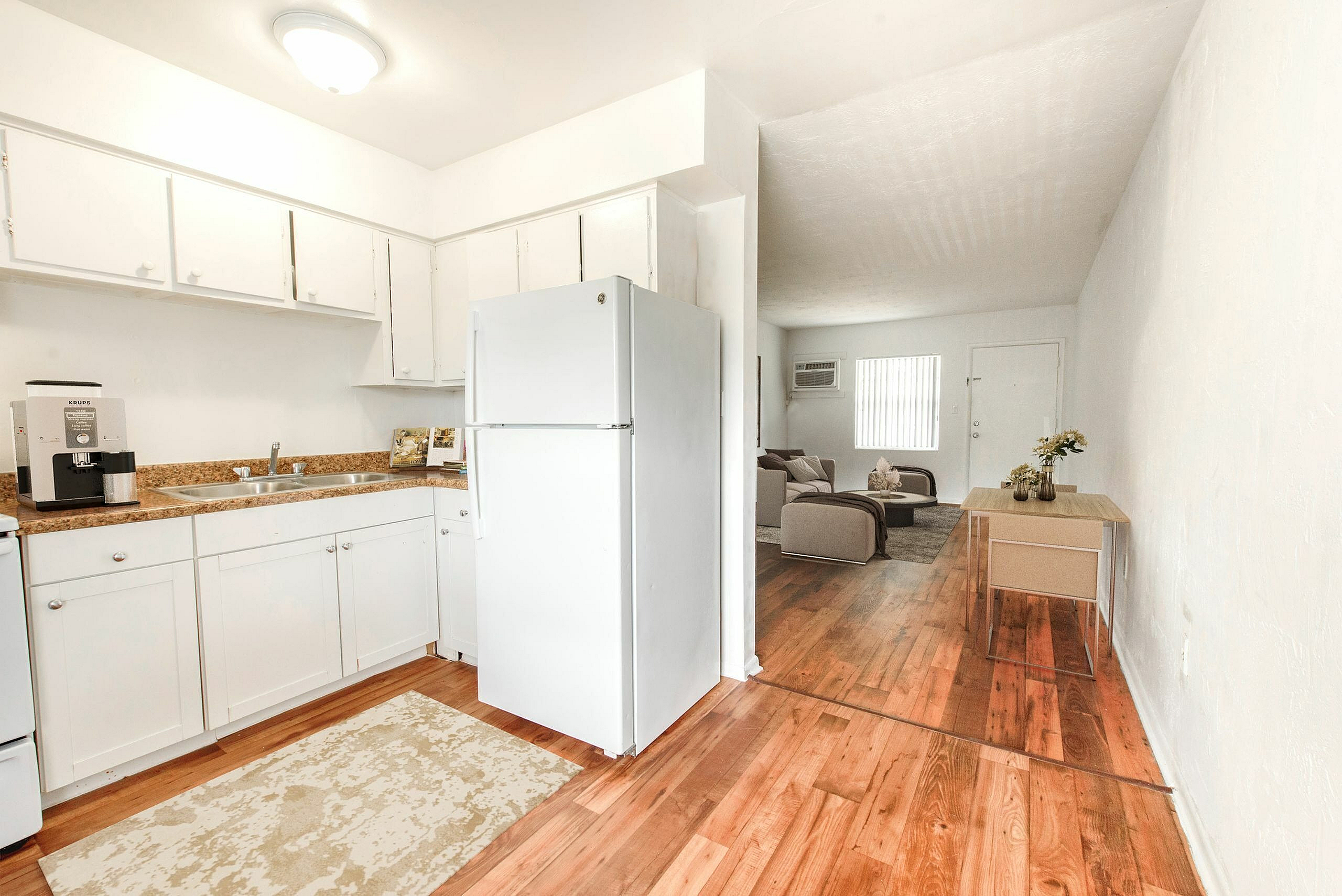 Bright and inviting apartment kitchen with sleek white cabinetry, modern appliances, and warm wooden flooring leading to a cozy living area.