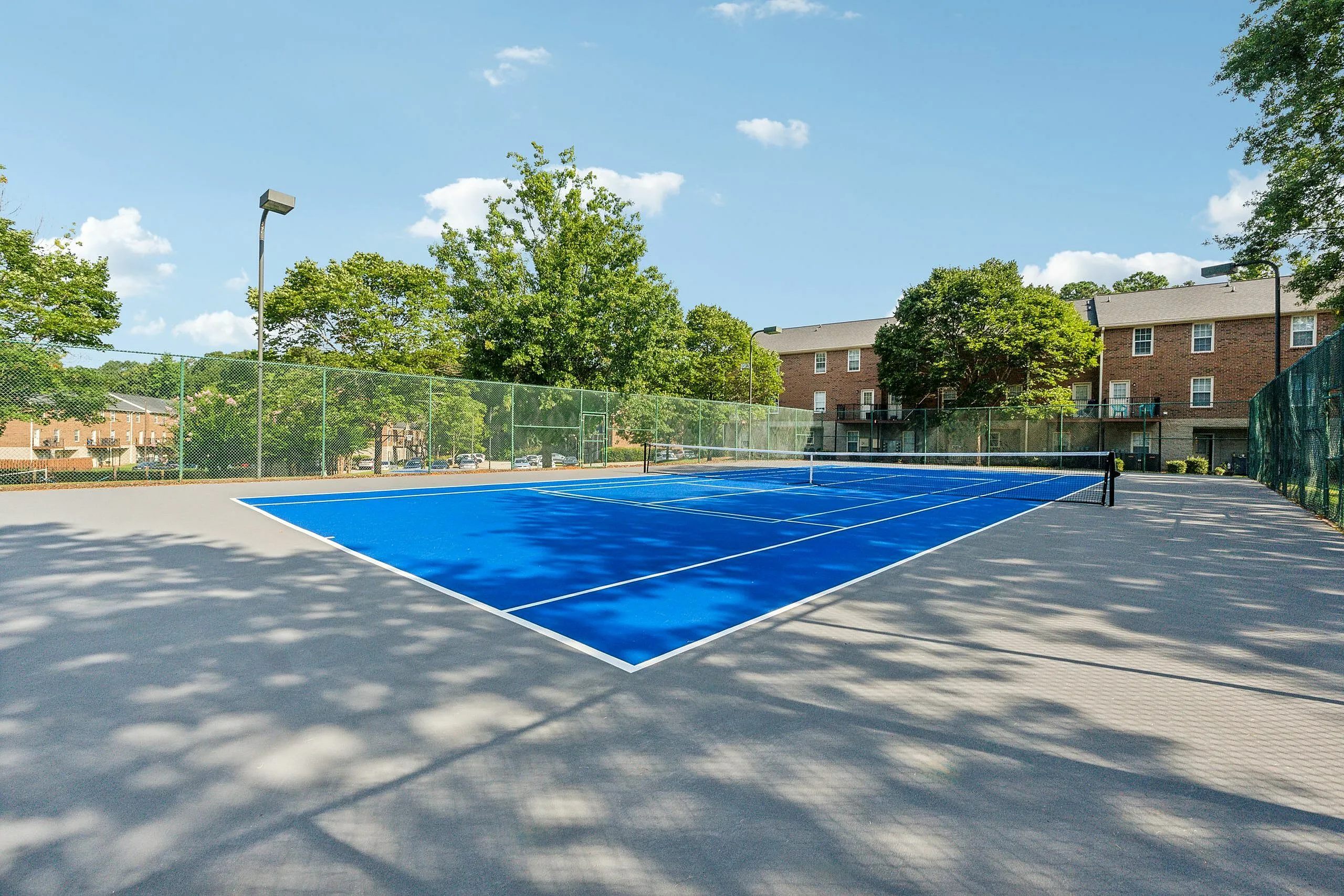 Vibrant blue tennis court surrounded by lush greenery and charming brick apartments, offering a perfect blend of recreation and community living.