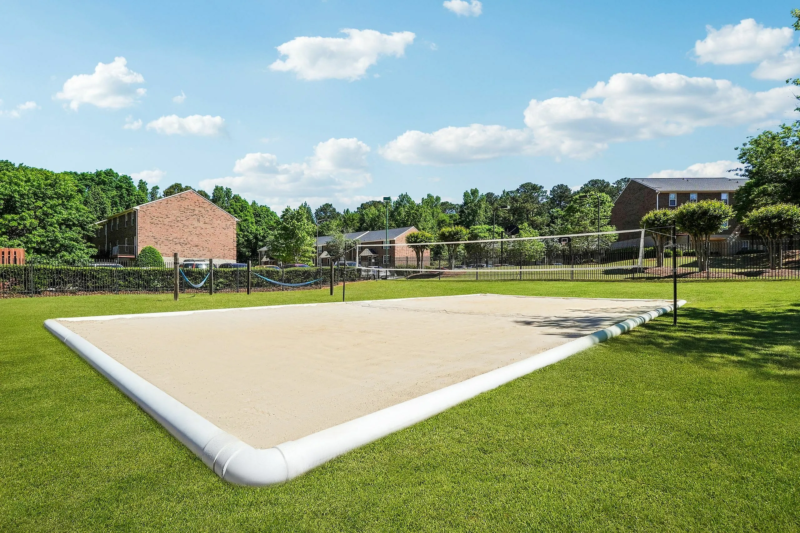 Inviting sand volleyball court surrounded by lush greenery and charming brick buildings under a clear blue sky.