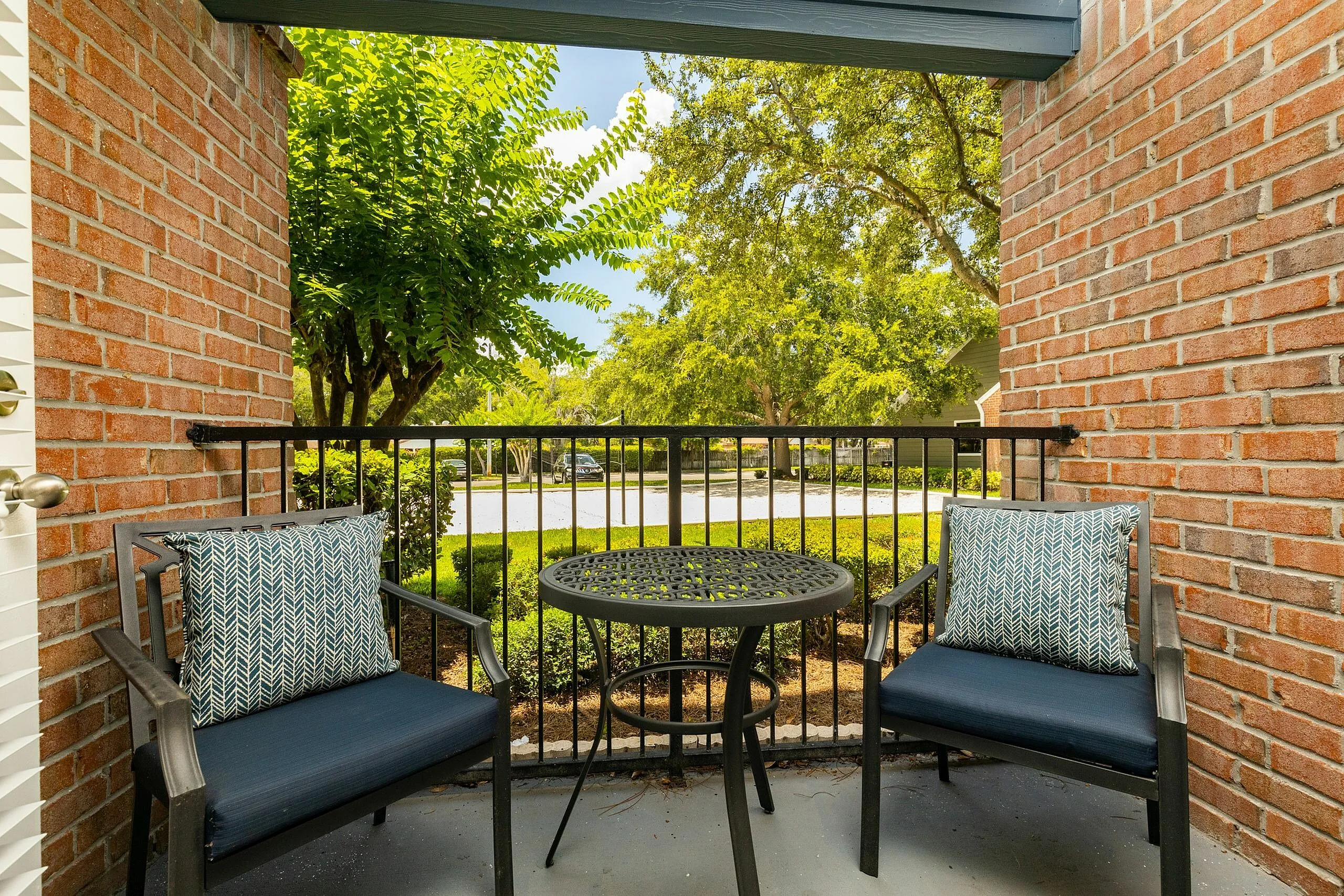 Charming balcony with cozy seating and a round table, surrounded by lush greenery and red brick walls, offering a serene outdoor retreat.