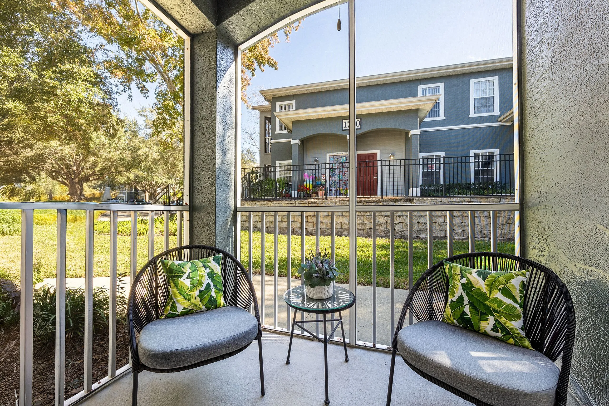Charming screened patio with cozy seating, perfect for relaxing. Overlooks lush greenery and a picturesque residential building.