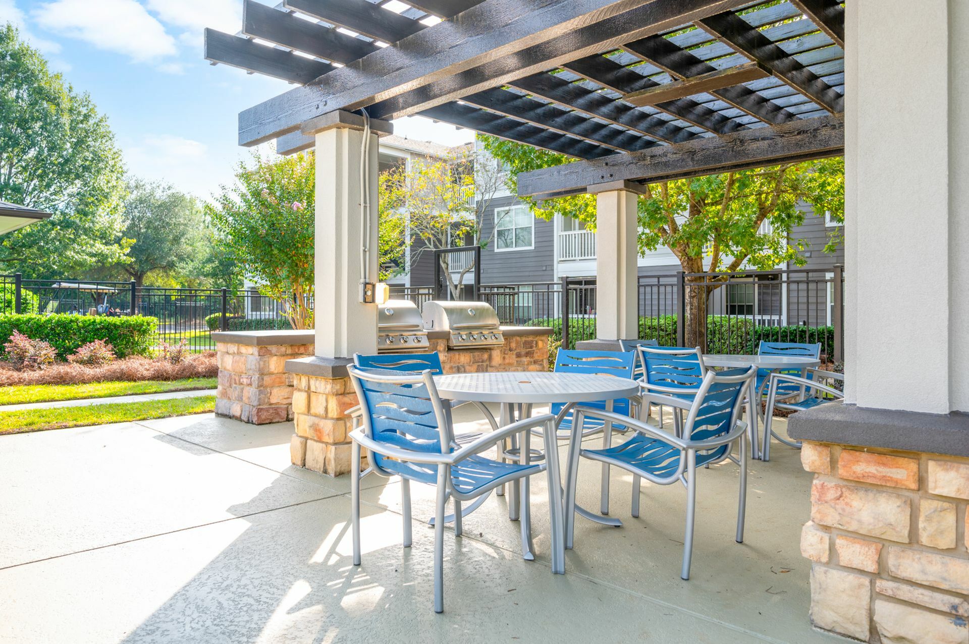 Inviting outdoor patio area with modern blue chairs and a stone counter featuring stainless steel barbecue grills, surrounded by lush greenery.