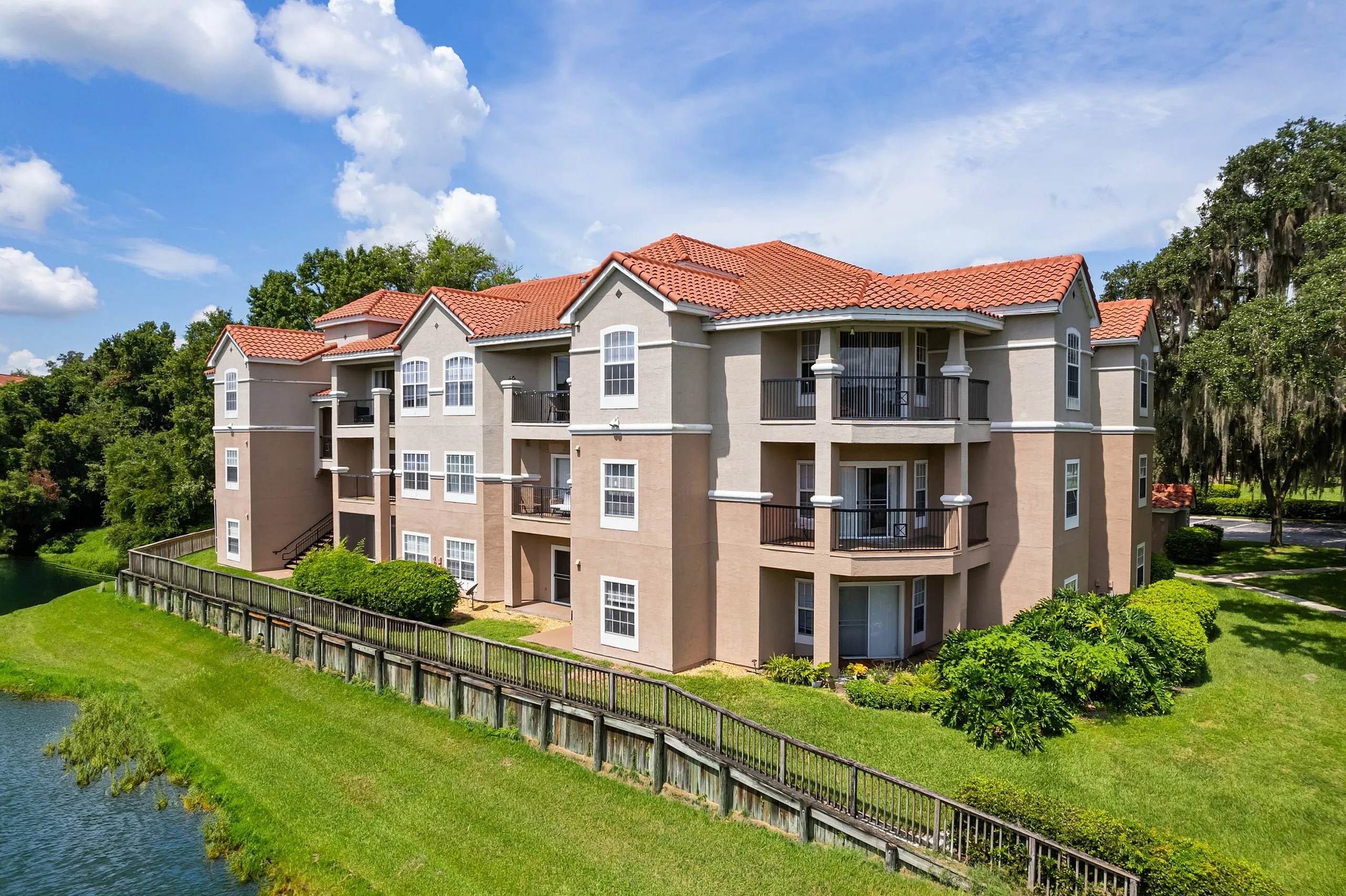 Charming multi-level apartment complex with elegant balconies, red-tiled roofs, lush landscaping, and a serene lakeside view under a bright, blue sky.