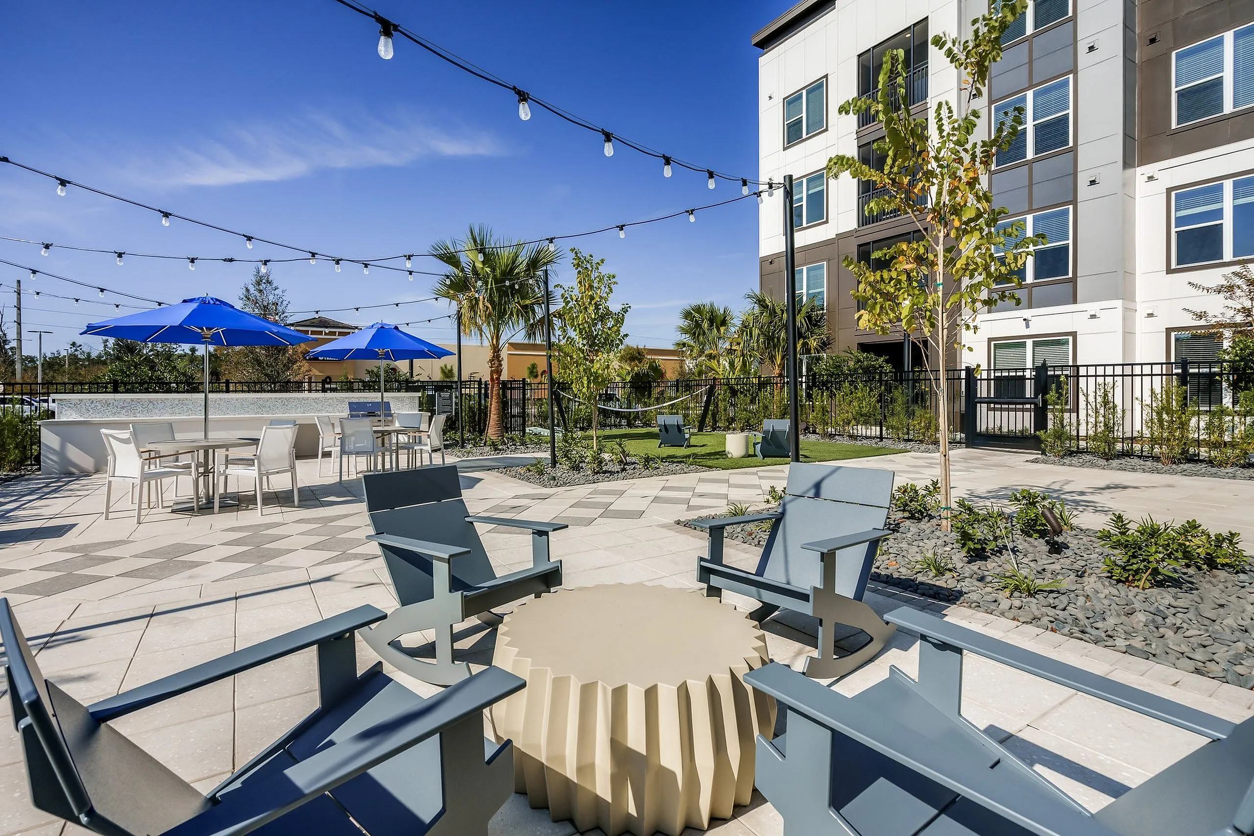 Modern apartment outdoor lounge area with sleek chairs, stylish stone flooring, vibrant blue umbrellas, and lush greenery, offering a luxurious and relaxing environment.