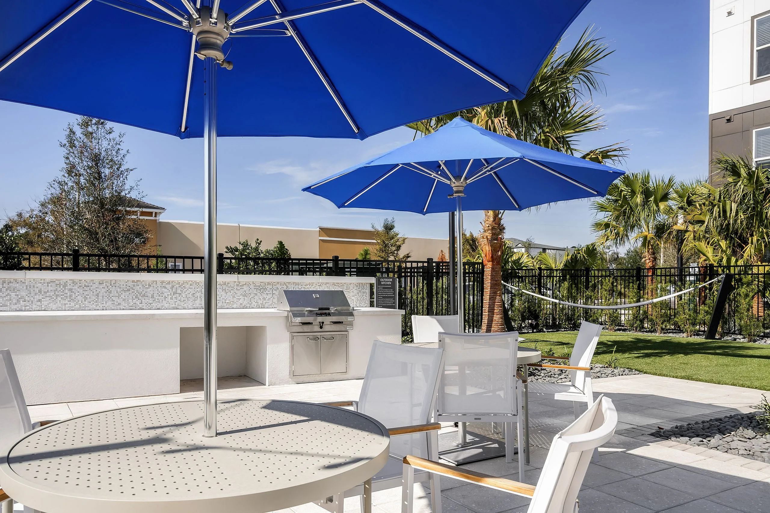 Chic outdoor patio area with blue umbrellas, stylish chairs, a modern barbecue grill, and lush greenery.
