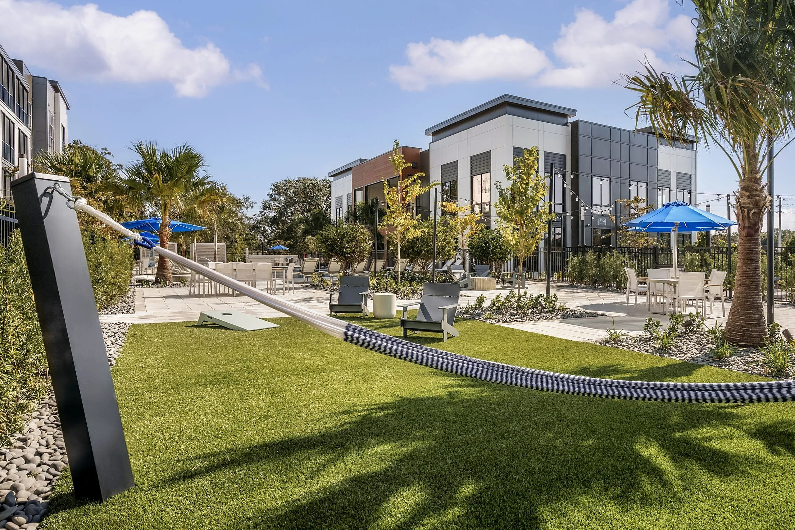 Modern apartment courtyard with lush green lawn, cozy hammock, stylish seating, and vibrant blue umbrellas under a bright sky.