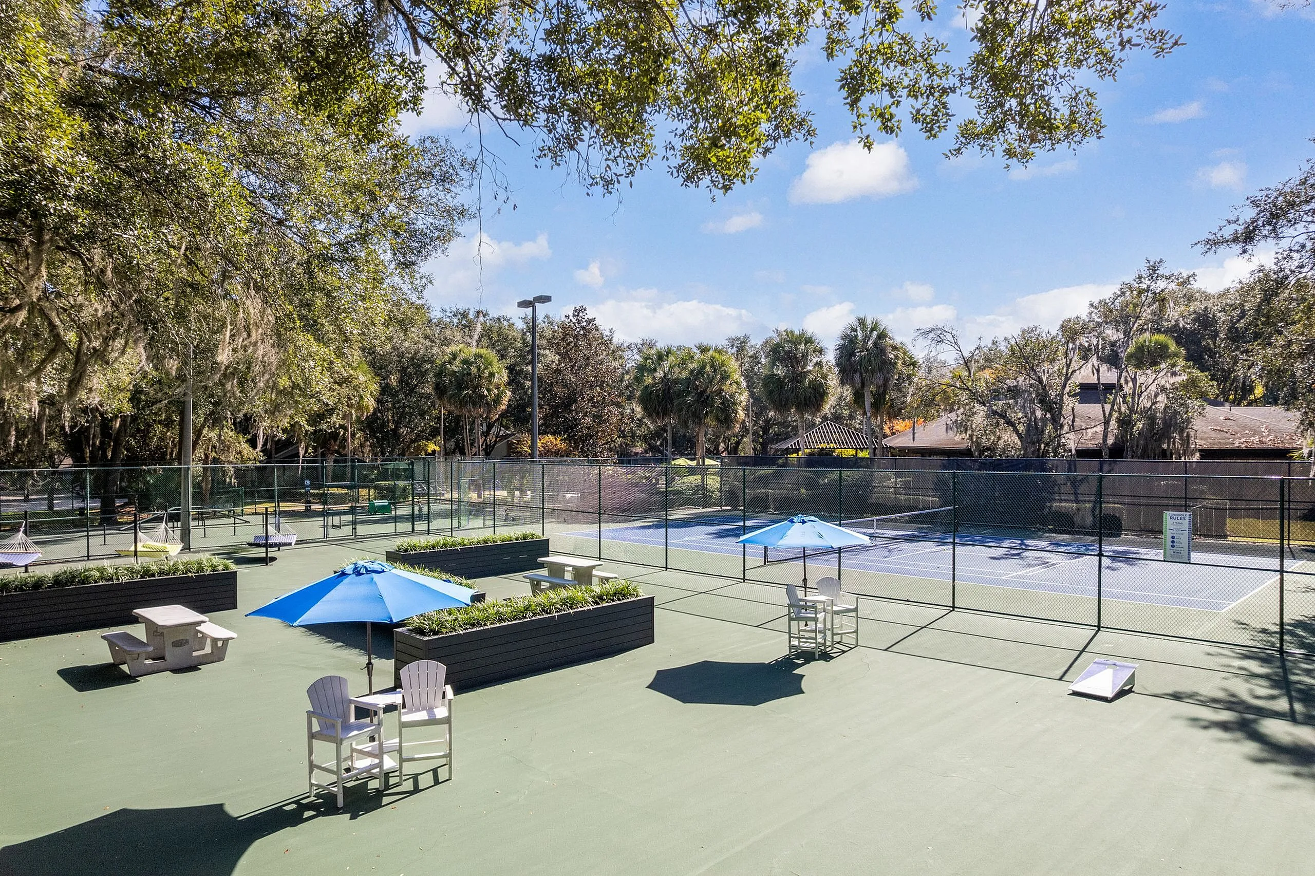 Inviting outdoor tennis court with nearby seating and vibrant blue umbrellas, surrounded by lush greenery for a relaxing atmosphere.
