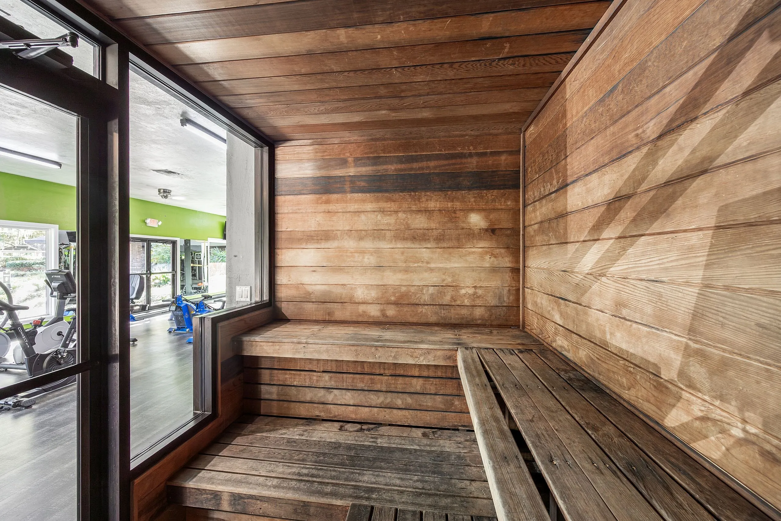Inviting wooden sauna with a modern glass door overlooking a well-equipped gym area.