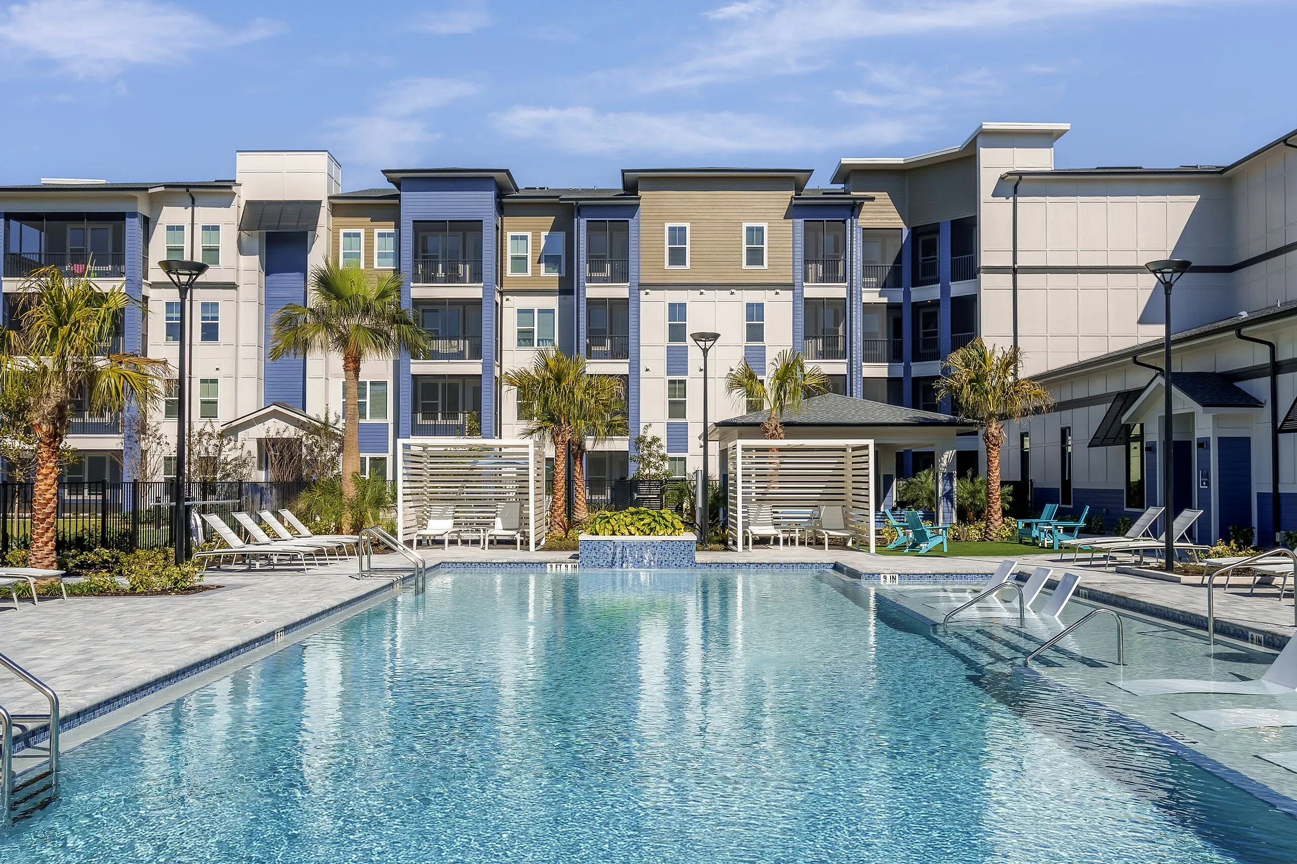 Luxurious apartment complex with a sparkling pool, surrounded by loungers and palm trees, under a clear blue sky.