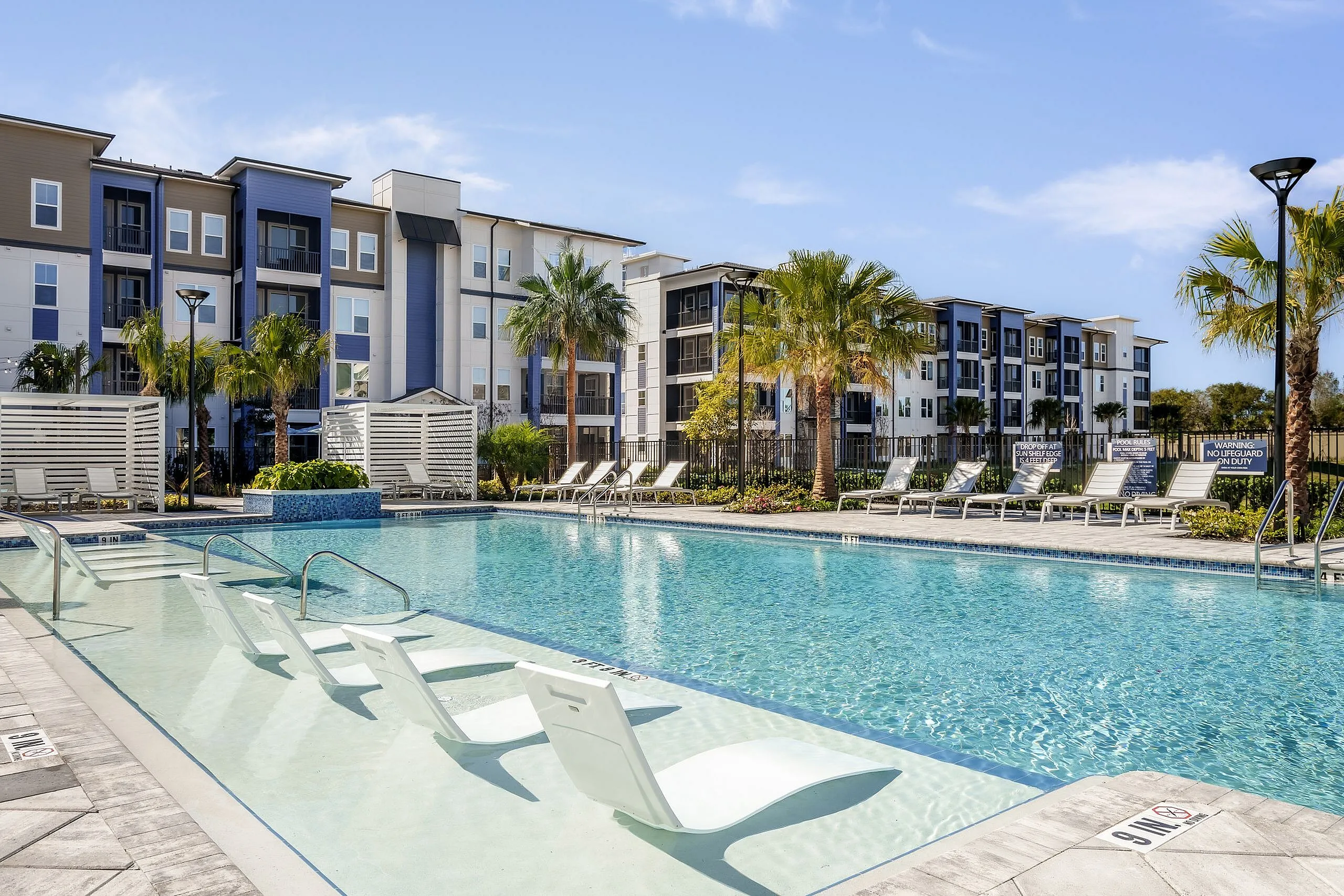 Luxurious apartment complex featuring a sparkling swimming pool surrounded by elegant lounge chairs and vibrant palm trees, perfect for relaxation and leisure.