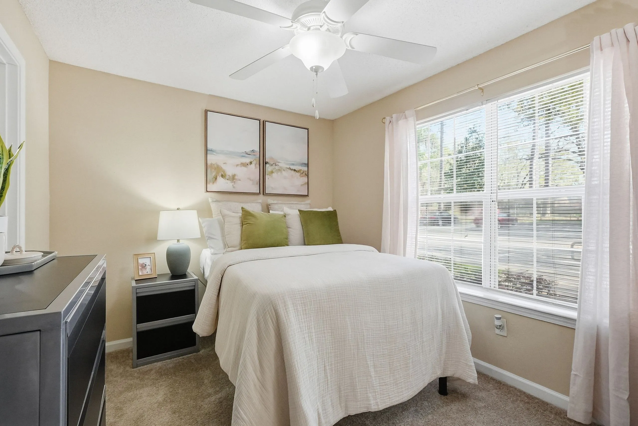 Inviting bedroom with soft beige walls, luxurious white bedding, and tranquil artwork, bathed in natural light from a large window, offering a peaceful retreat.