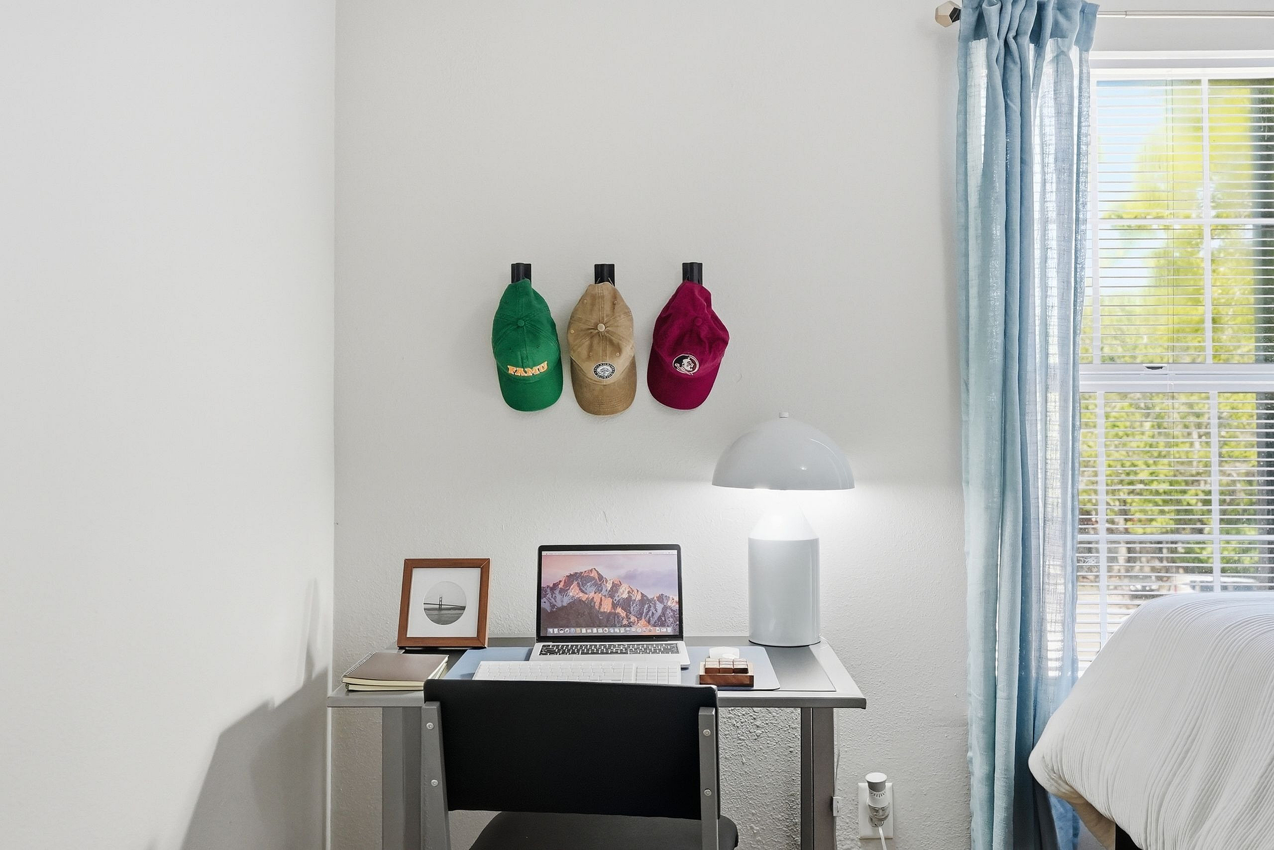 Modern workspace in bright apartment bedroom featuring a sleek desk with a laptop, contemporary lamp, and decor accents beneath colorful hats. Sunlit window with soft blue curtains adds a cozy touch.