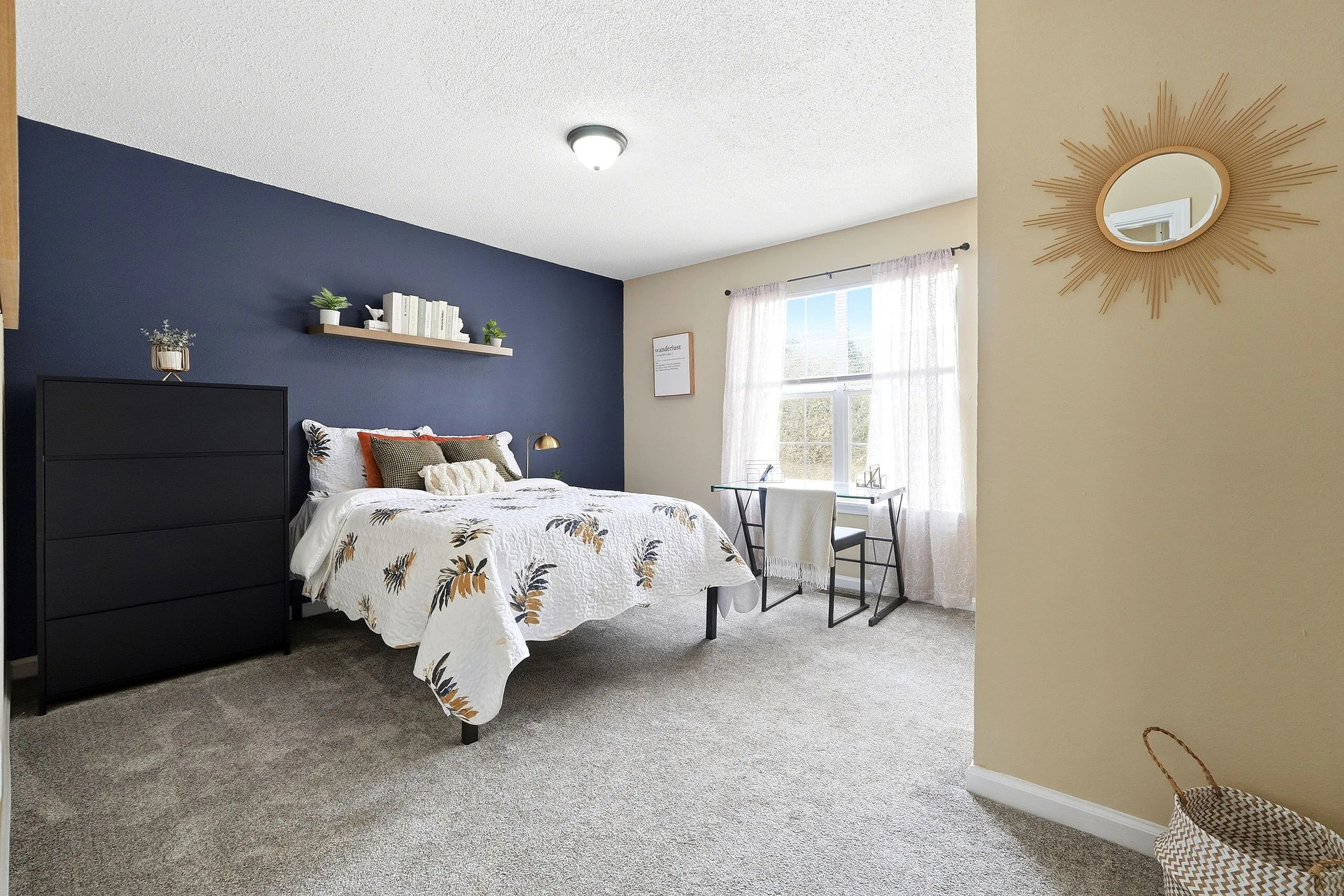Charming bedroom with stylish dark accent wall, cozy floral quilt, and sunburst mirror, featuring a bright window and elegant decor.