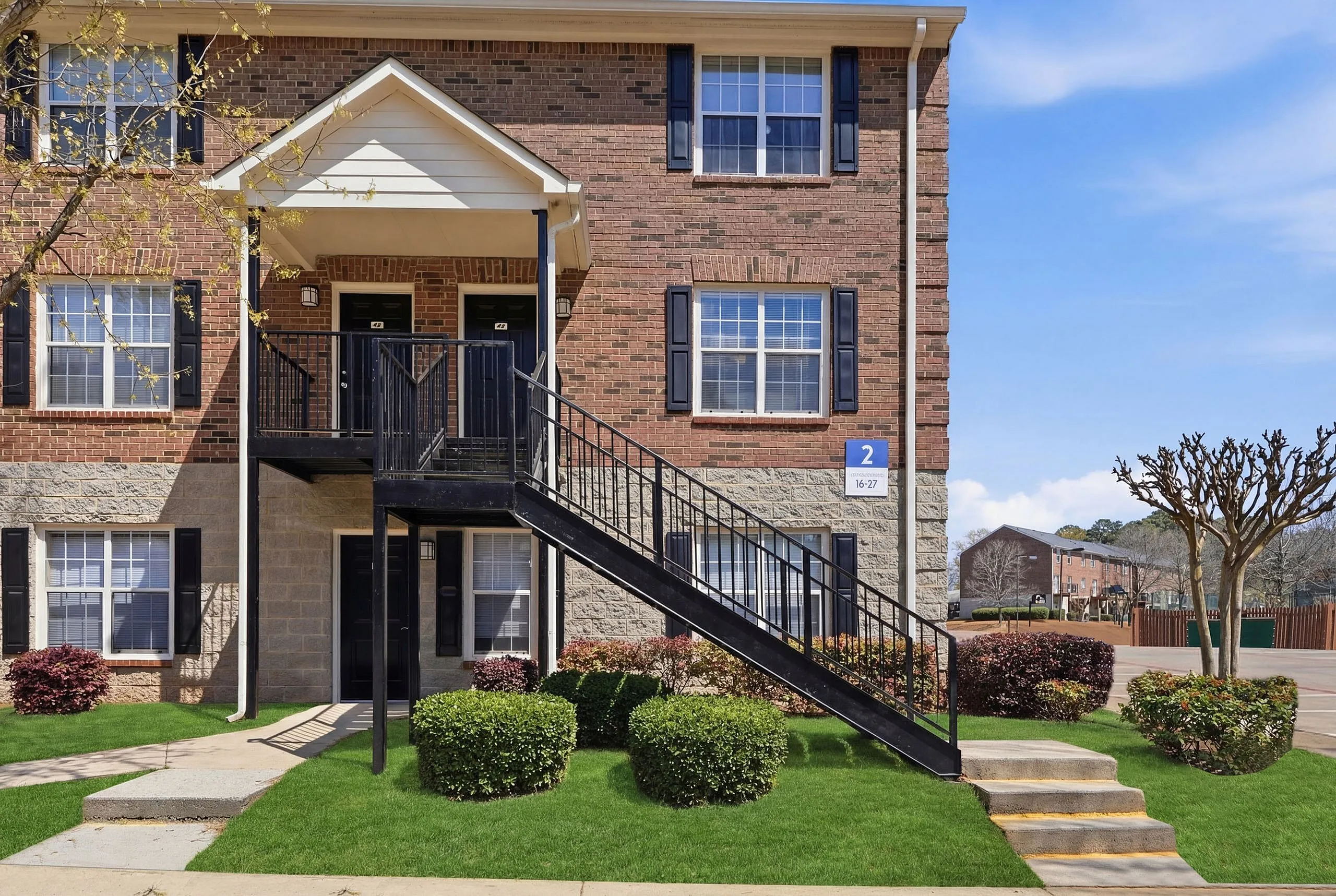 Charming brick apartment exterior with black stairs, featuring lush green landscaping and modern windows in a serene neighborhood.