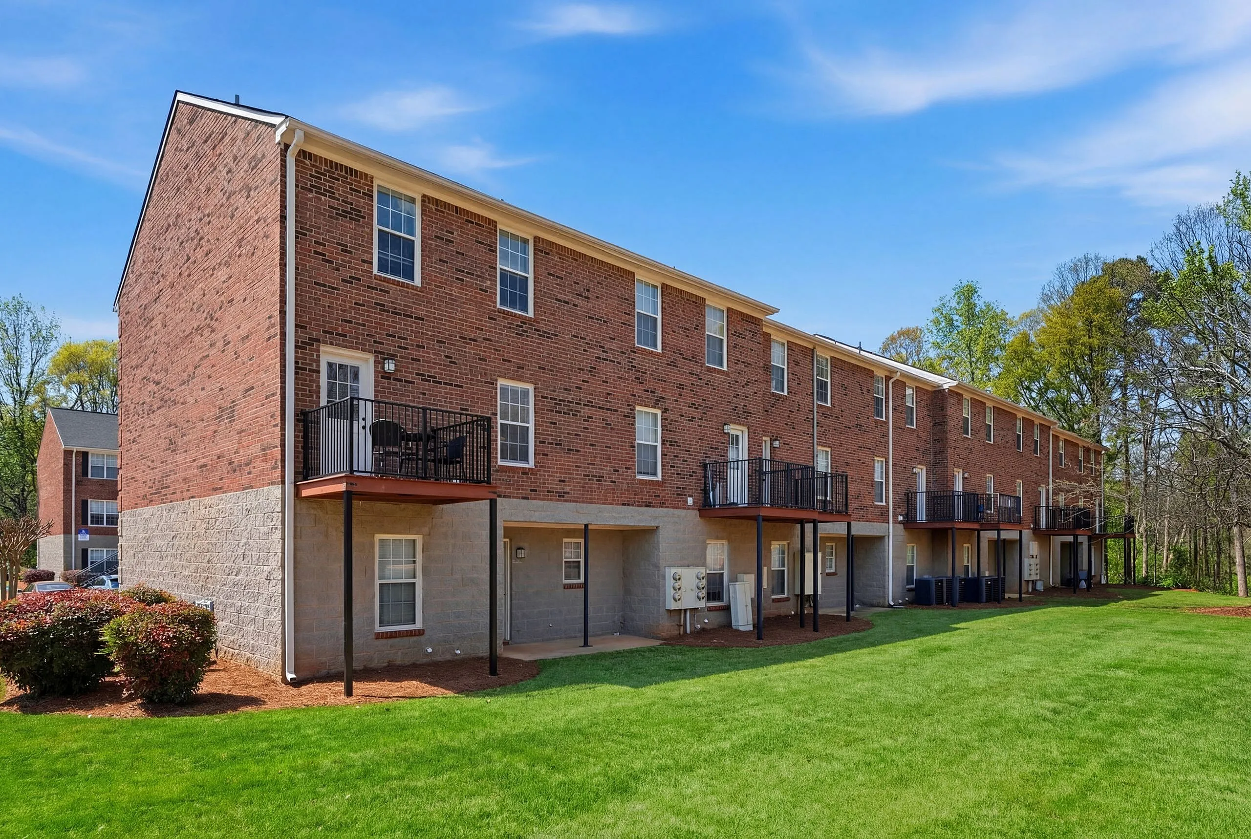 Charming brick apartment building with individual balconies, surrounded by lush green lawns and scenic trees, perfect for serene living.
