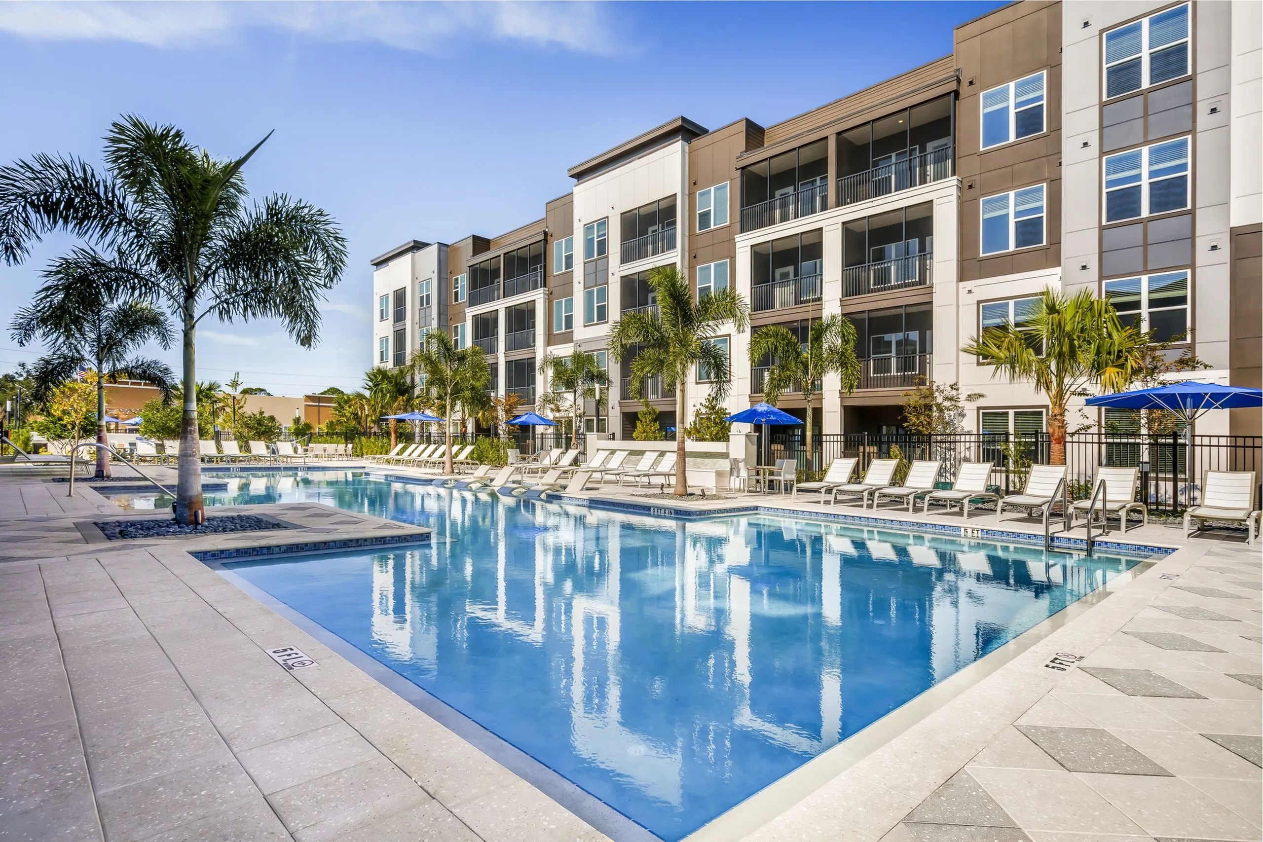 Modern apartment complex featuring a sparkling pool, surrounded by palm trees and stylish lounge chairs under blue umbrellas, offering a luxurious living experience.