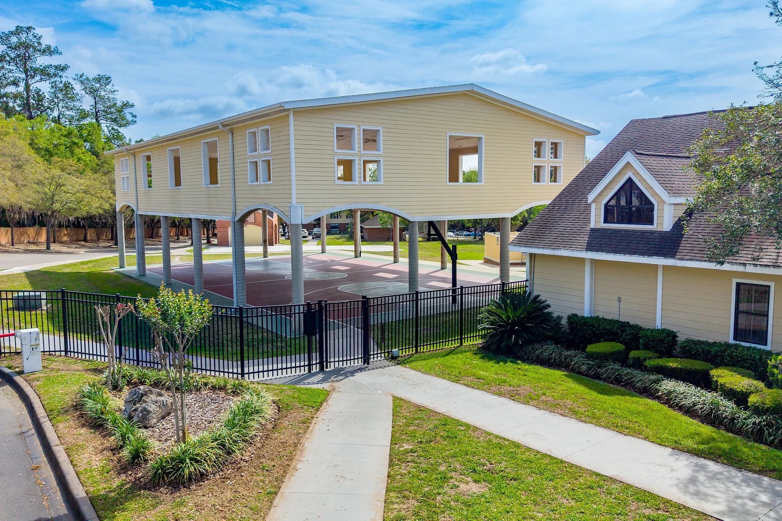 Elevated modern architecture with arched supports above a basketball court, surrounded by lush greenery and elegant fencing.