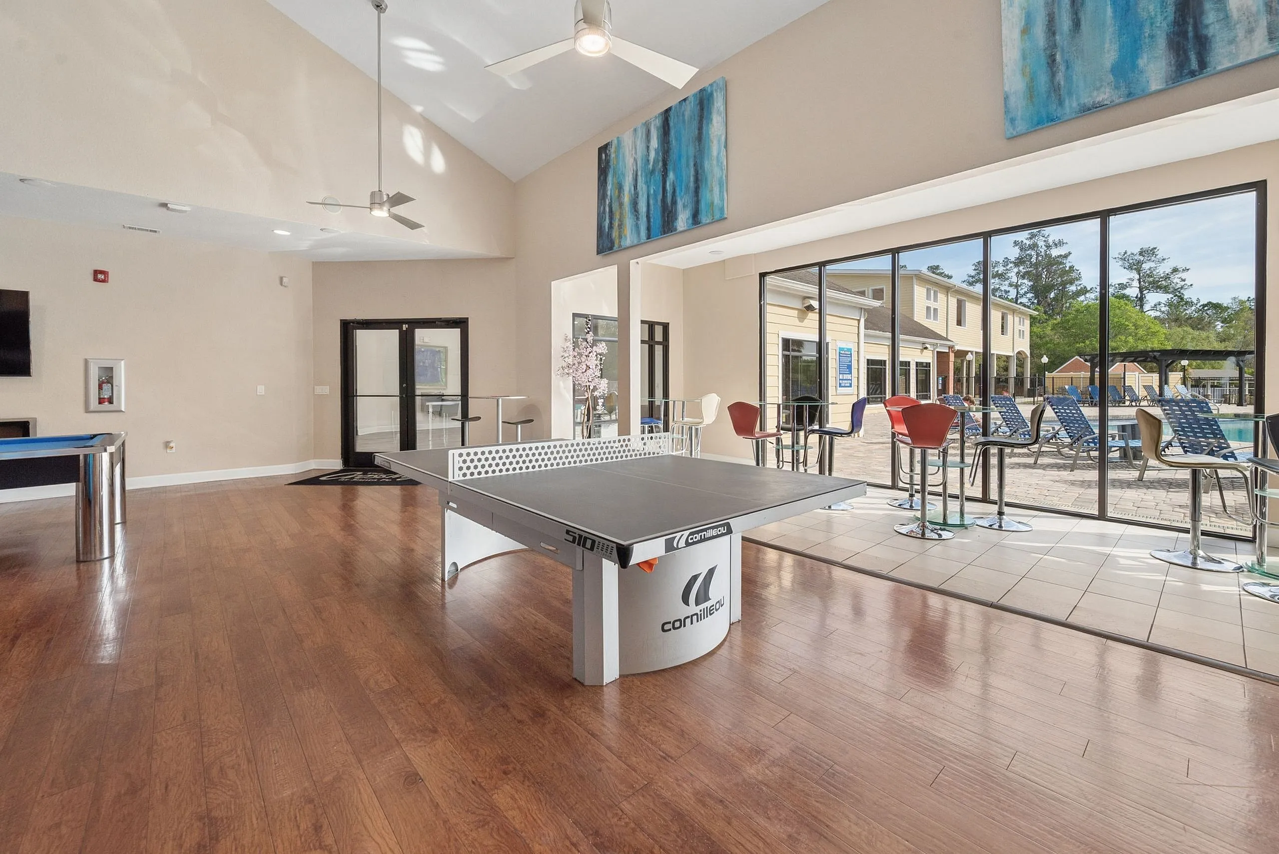 Modern recreation area with a sleek ping pong table, stylish high chairs, and large windows overlooking a sunny patio with poolside loungers.