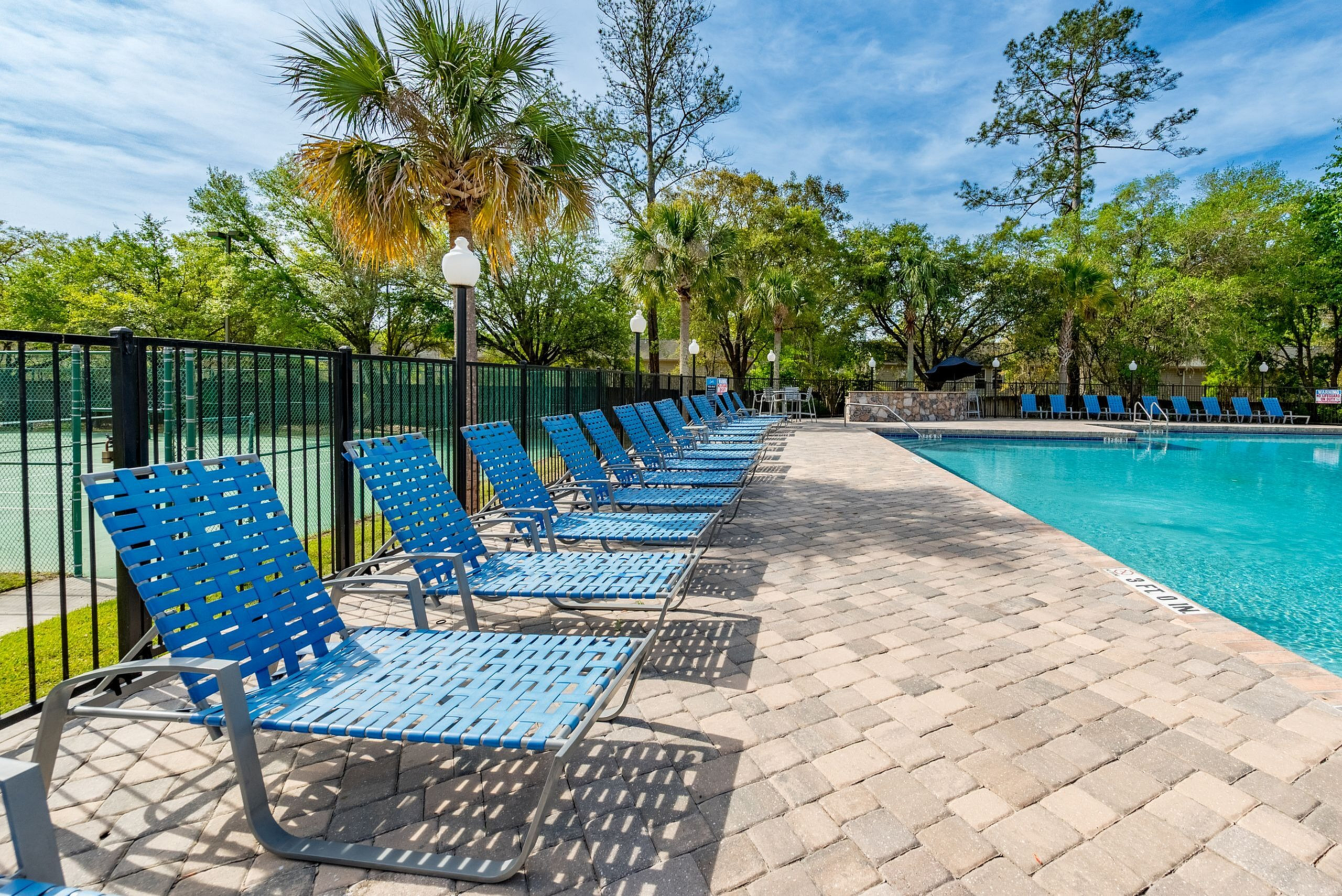 Inviting swimming pool area with stylish blue lounge chairs, surrounded by lush green trees and a bright blue sky.