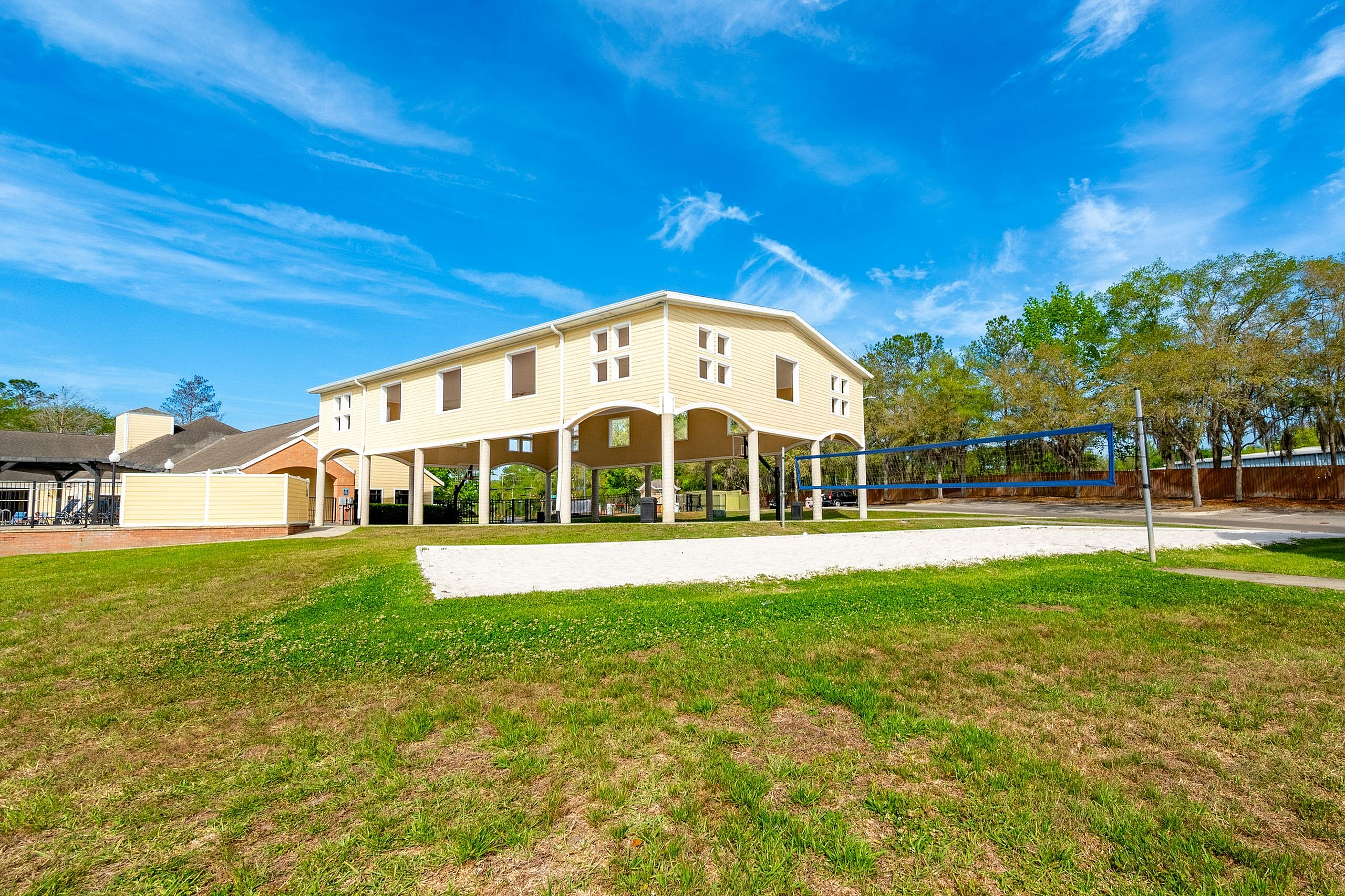 Bright and inviting exterior of a property featuring an elevated structure and a well-maintained sand volleyball court, surrounded by lush greenery under a clear blue sky.
