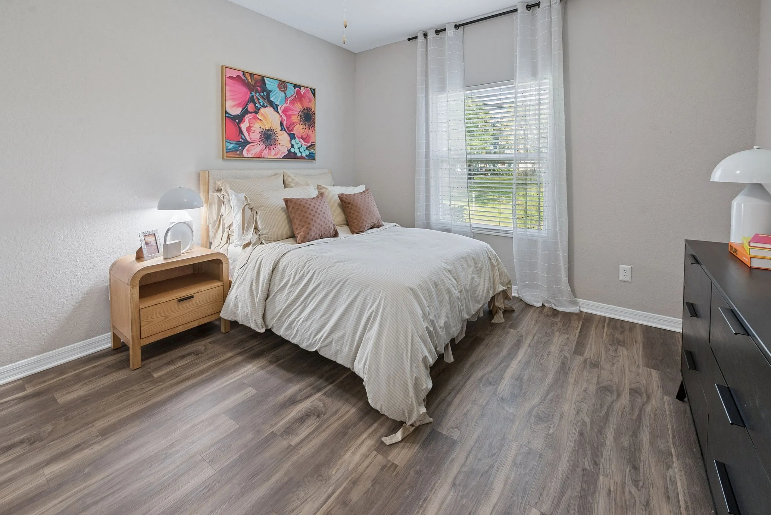Bright and inviting bedroom with elegant hardwood floors, featuring a cozy bed, vibrant floral artwork, and abundant natural light streaming through sheer curtains.