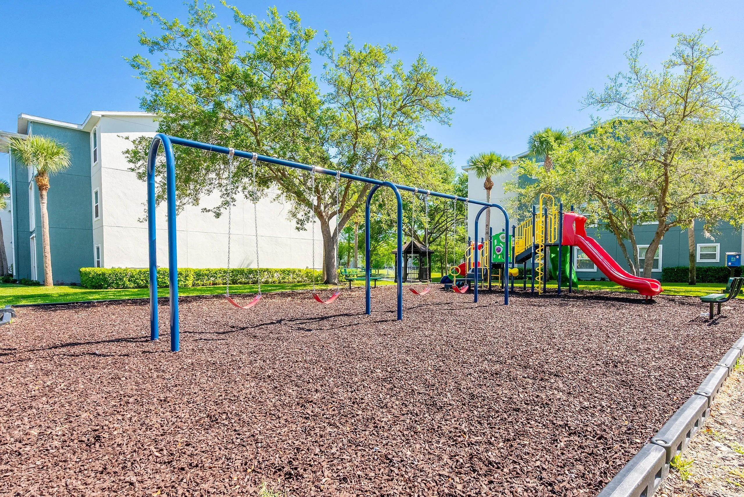 Vibrant playground in a lush residential area, featuring swings and a colorful slide set against a backdrop of green trees and modern apartments.
