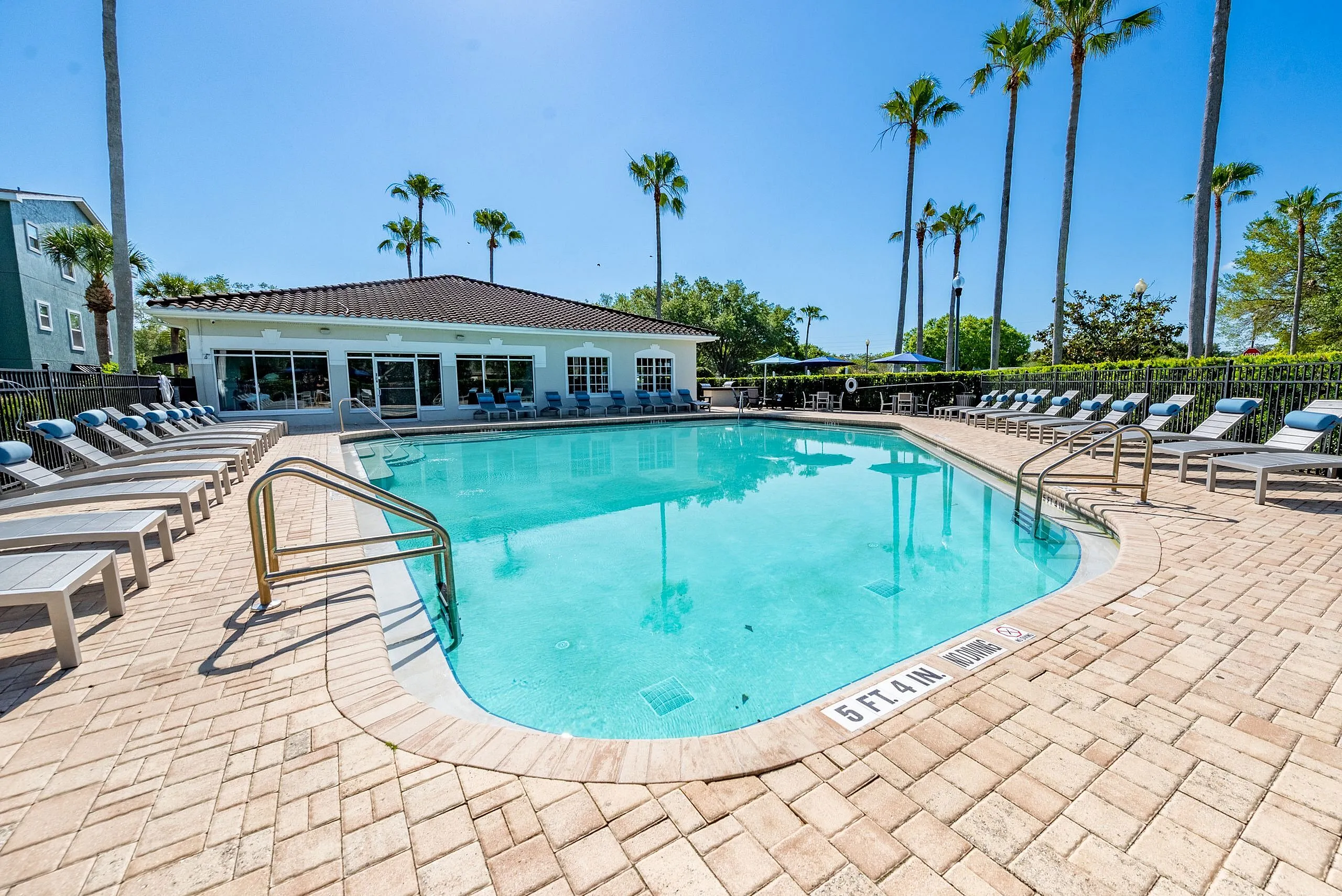 Inviting outdoor pool area with sun loungers, surrounded by lush palm trees and a modern clubhouse, perfect for relaxation.