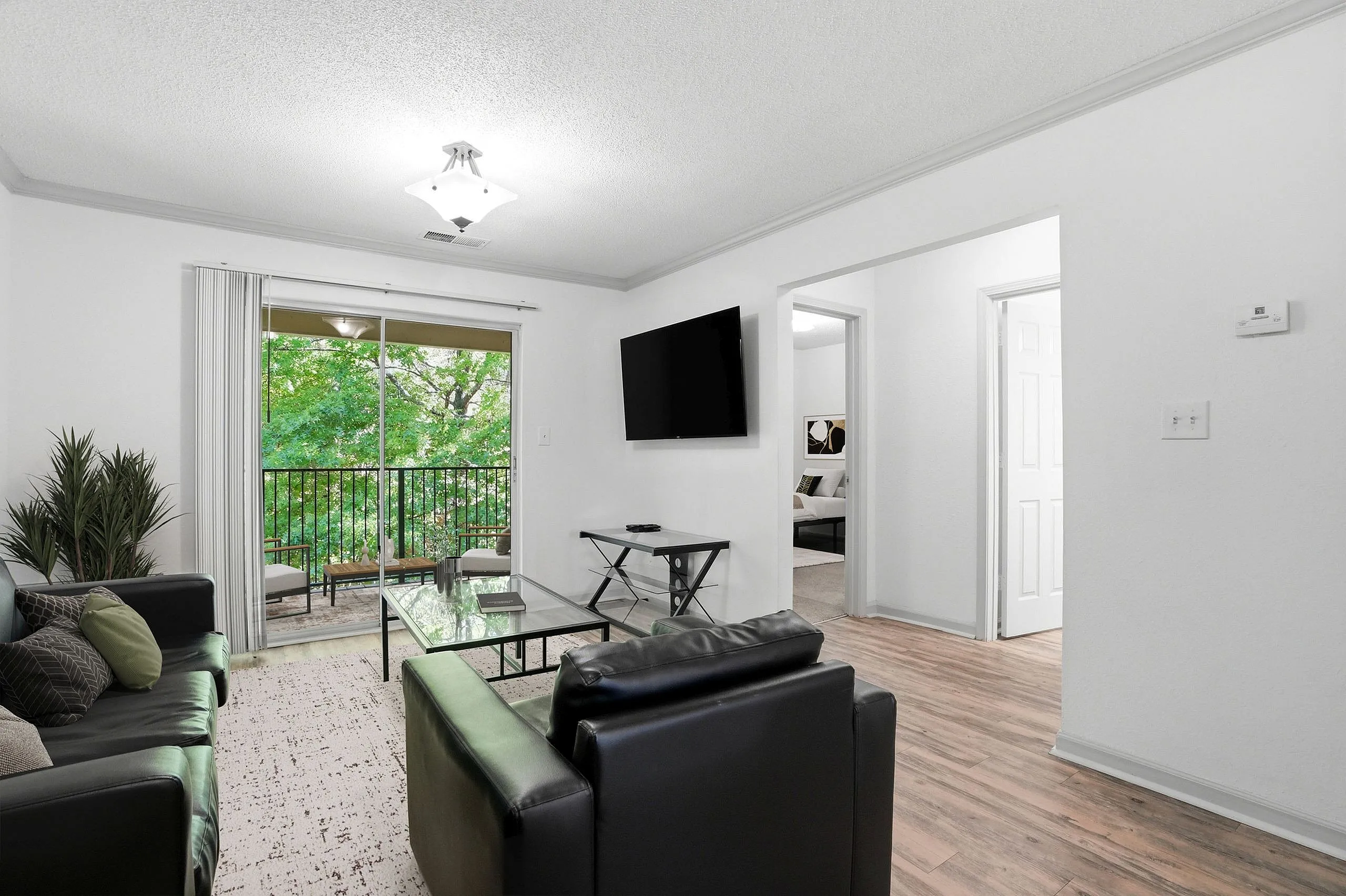Elegant living room with modern black leather sofas, a flat screen TV, and a view of lush green trees through large glass doors leading to a balcony.