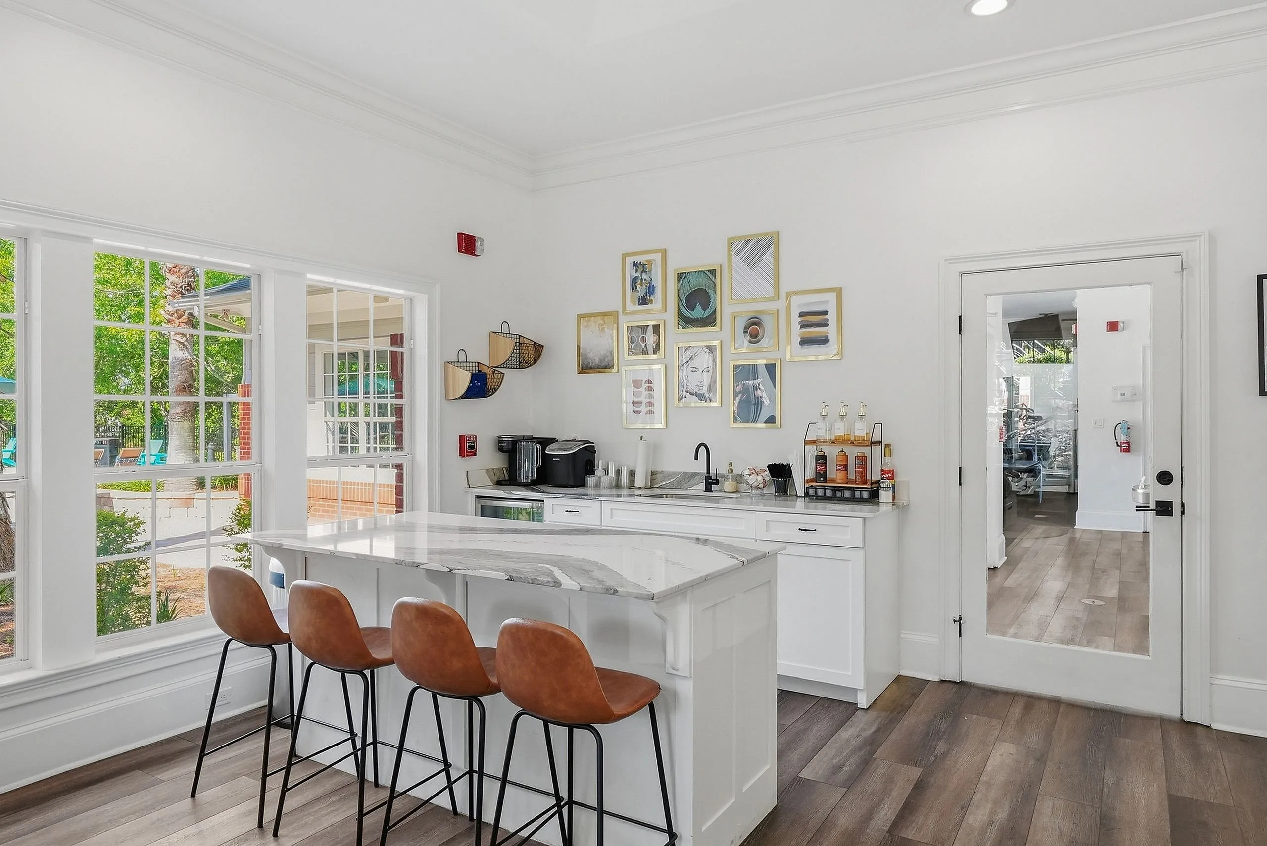 Chic and modern kitchen space with a marble island, leather stools, and a wall adorned with stylish artwork, set against large windows offering a bright and airy ambiance.
