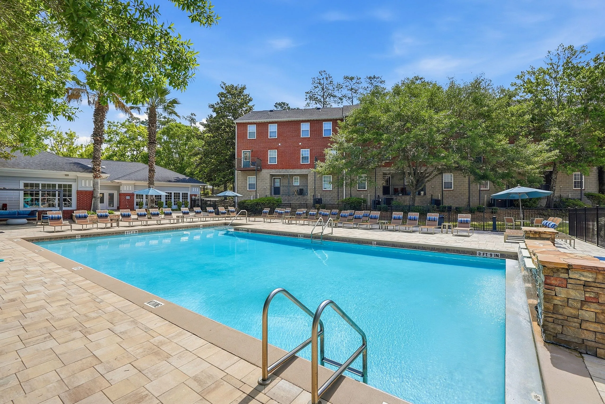 Inviting outdoor pool area with sun loungers, surrounded by lush greenery and charming brick buildings under a clear blue sky.