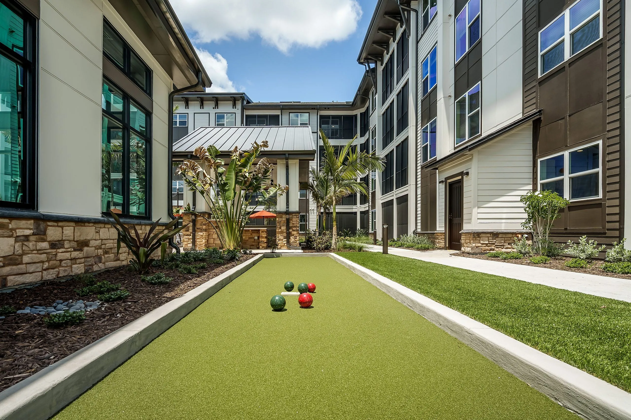 Modern apartment complex exterior with a lush landscaped bocce ball court, sleek architecture, and vibrant greenery.