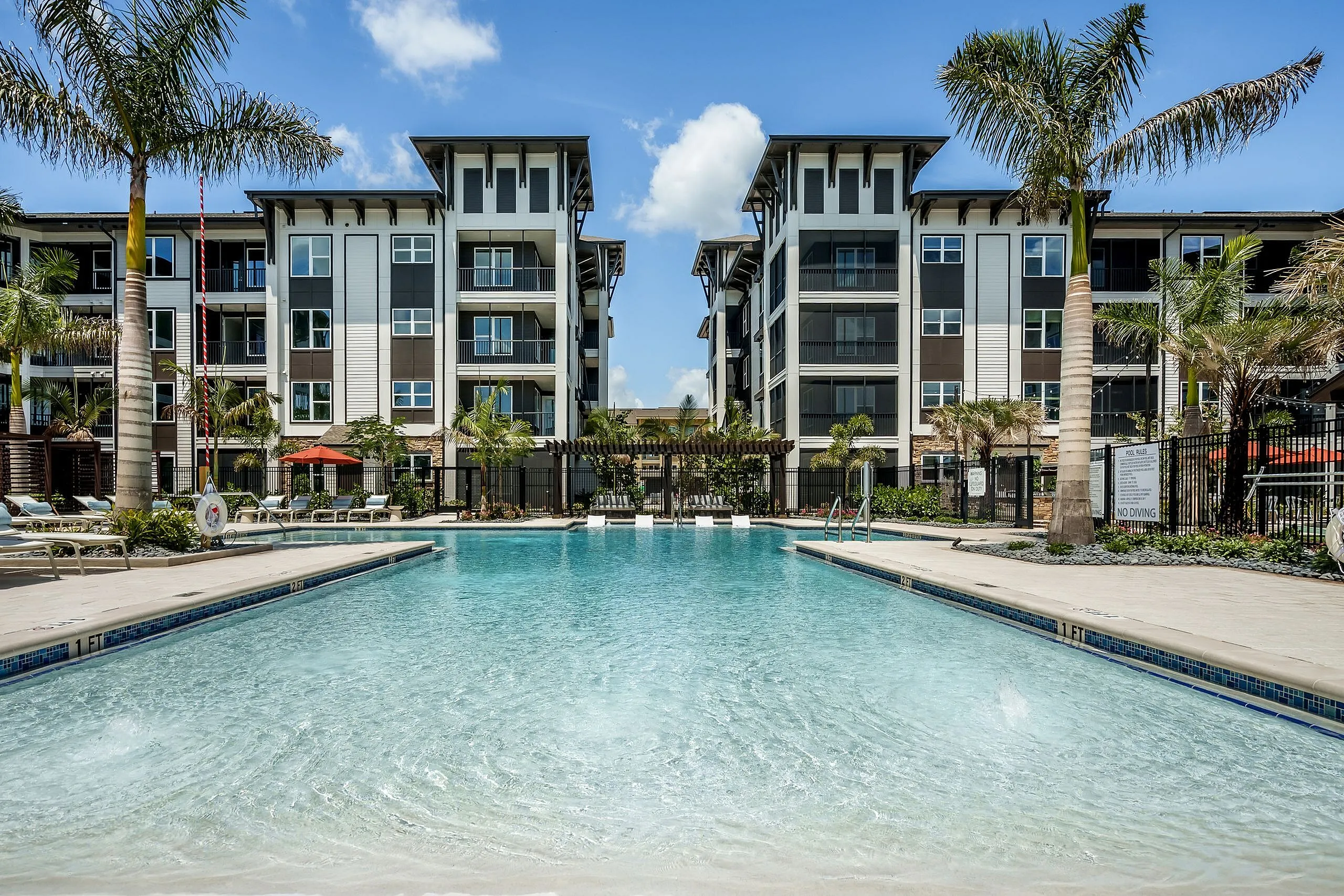 Luxurious apartment complex featuring a sparkling swimming pool, palm trees, and modern multi-story buildings.