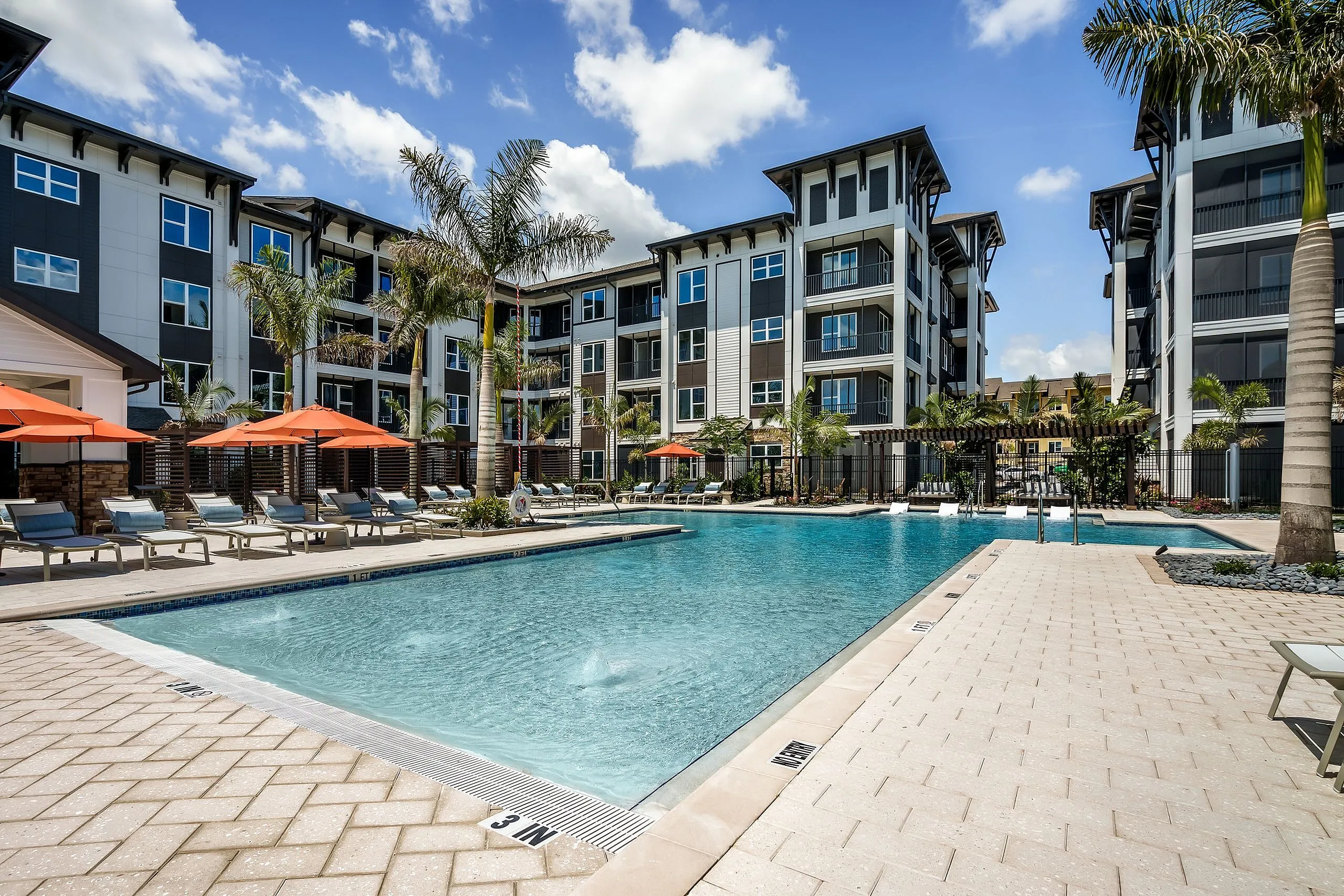 Luxurious apartment complex featuring a shimmering resort-style pool, elegant palm trees, and stylish sun loungers under vibrant orange umbrellas.