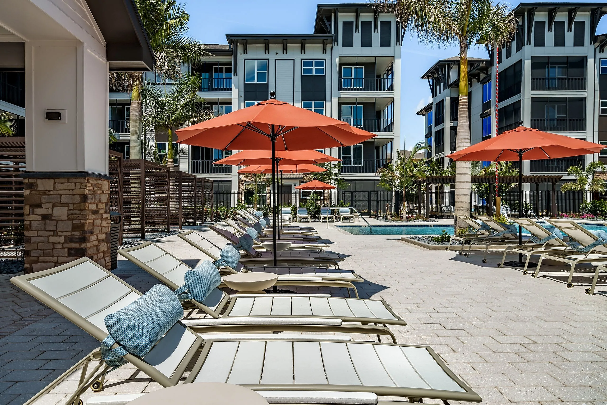 Luxurious apartment complex with vibrant outdoor pool area, sun loungers, and bright orange umbrellas surrounded by lush palm trees.