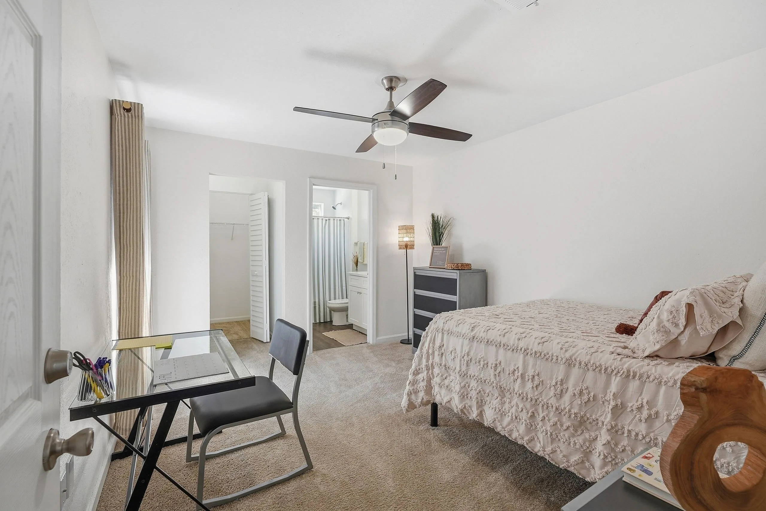 Bright and inviting bedroom with plush carpeting, a modern ceiling fan, and an en-suite bathroom. Includes a sleek glass desk for productive workspace.