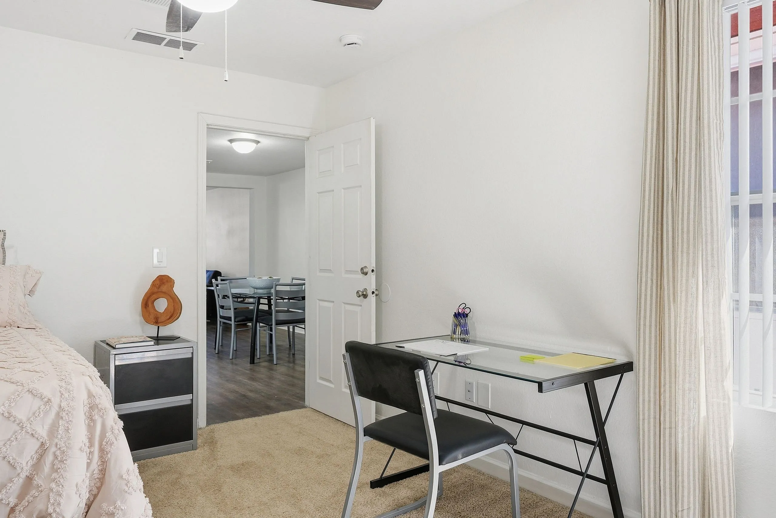 Bright, cozy bedroom with a modern glass desk and an open doorway leading to a stylish dining area.