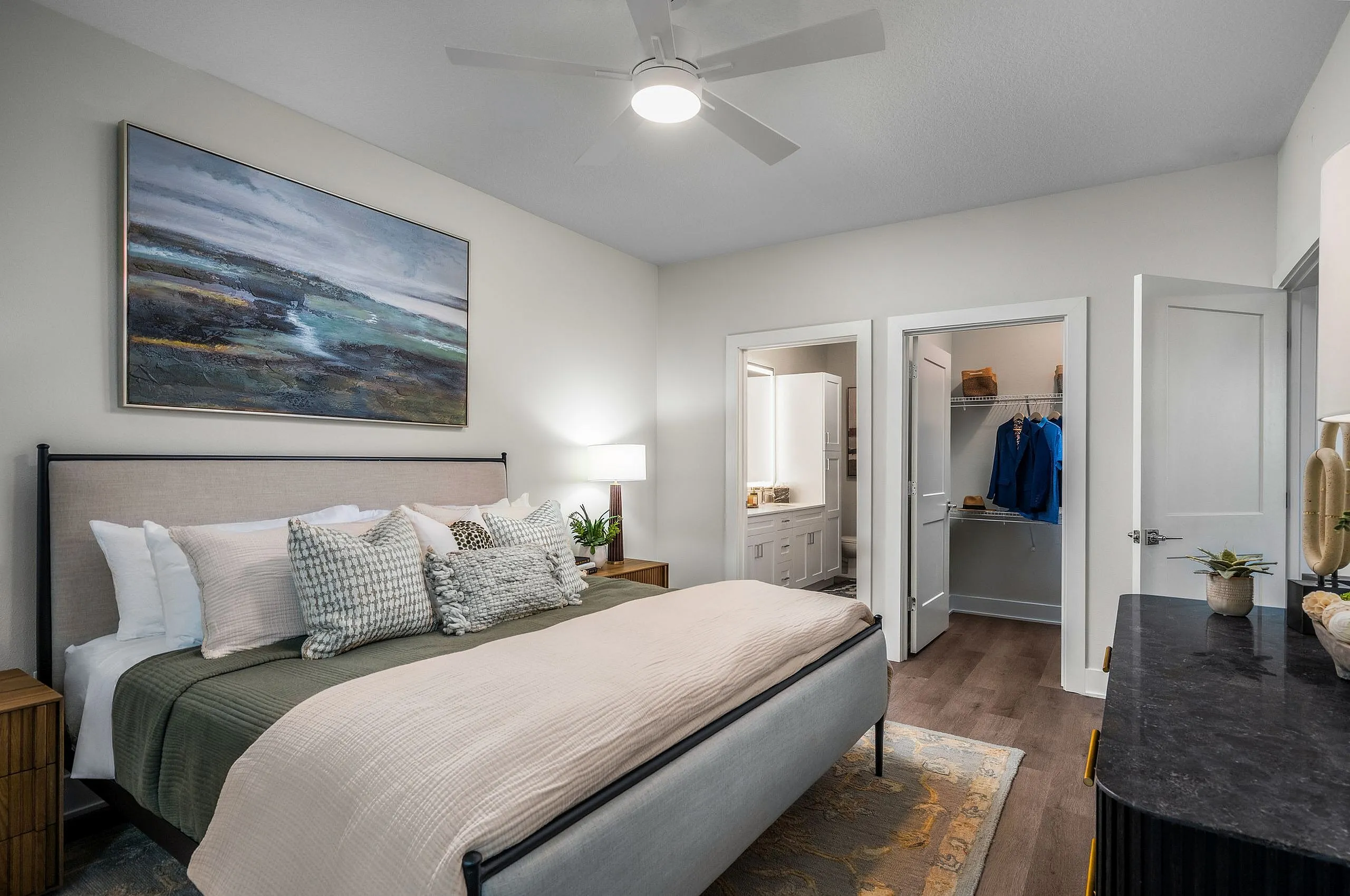 Elegant bedroom with stylish decor, featuring a plush bed, modern ceiling fan, and artful wall painting, adjacent to a well-organized walk-in closet and sleek en-suite bathroom.