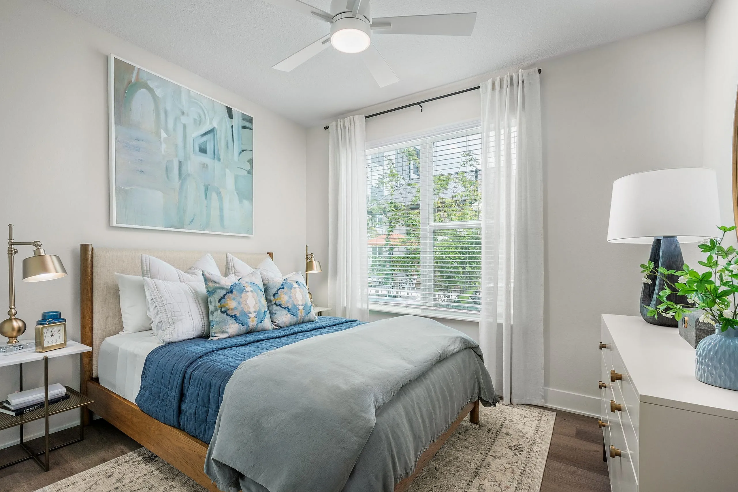 Bright and inviting bedroom with large window, modern ceiling fan, and elegant decor. Plush bedding and stylish artwork create a serene atmosphere.