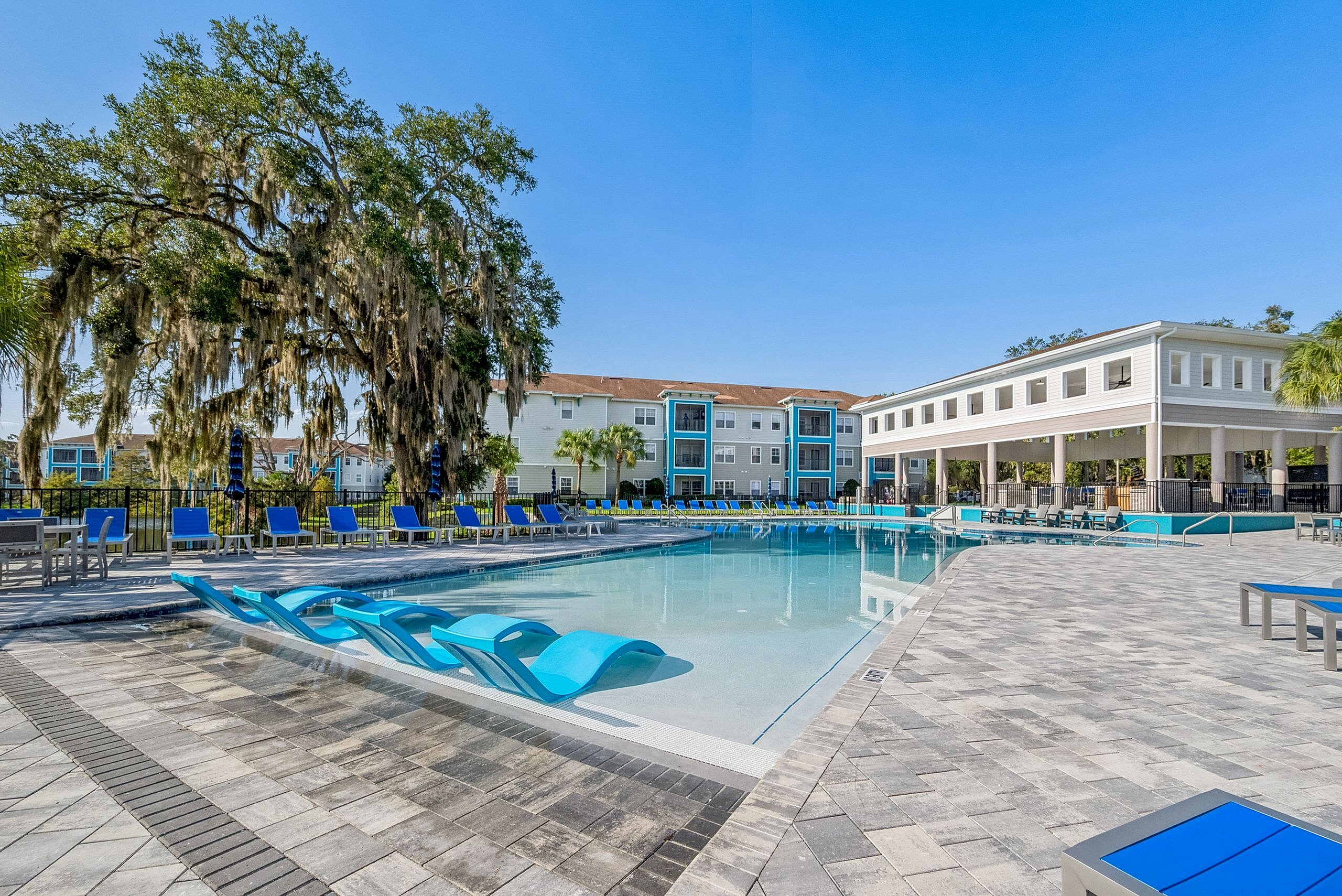 Luxurious apartment complex with a sparkling pool, spacious lounge chairs, and scenic views of lush greenery and modern architecture.