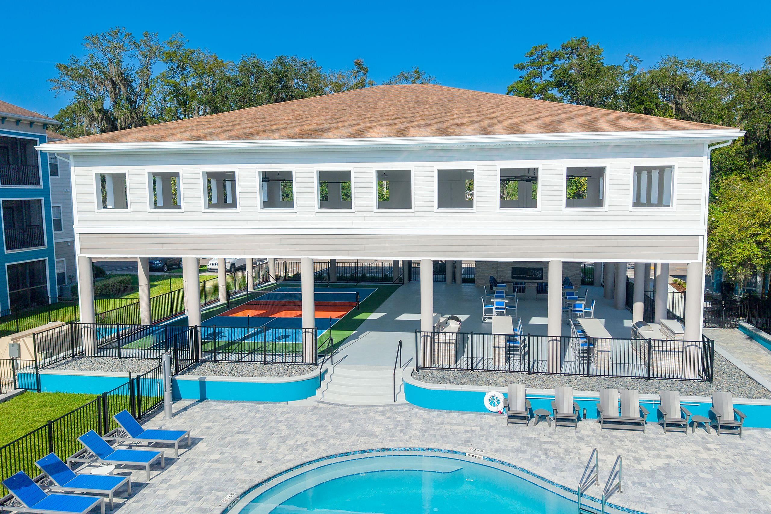 Modern apartment complex with a scenic outdoor space featuring a pristine pool, sun loungers, and multi-use sports courts, perfect for relaxation and recreation.