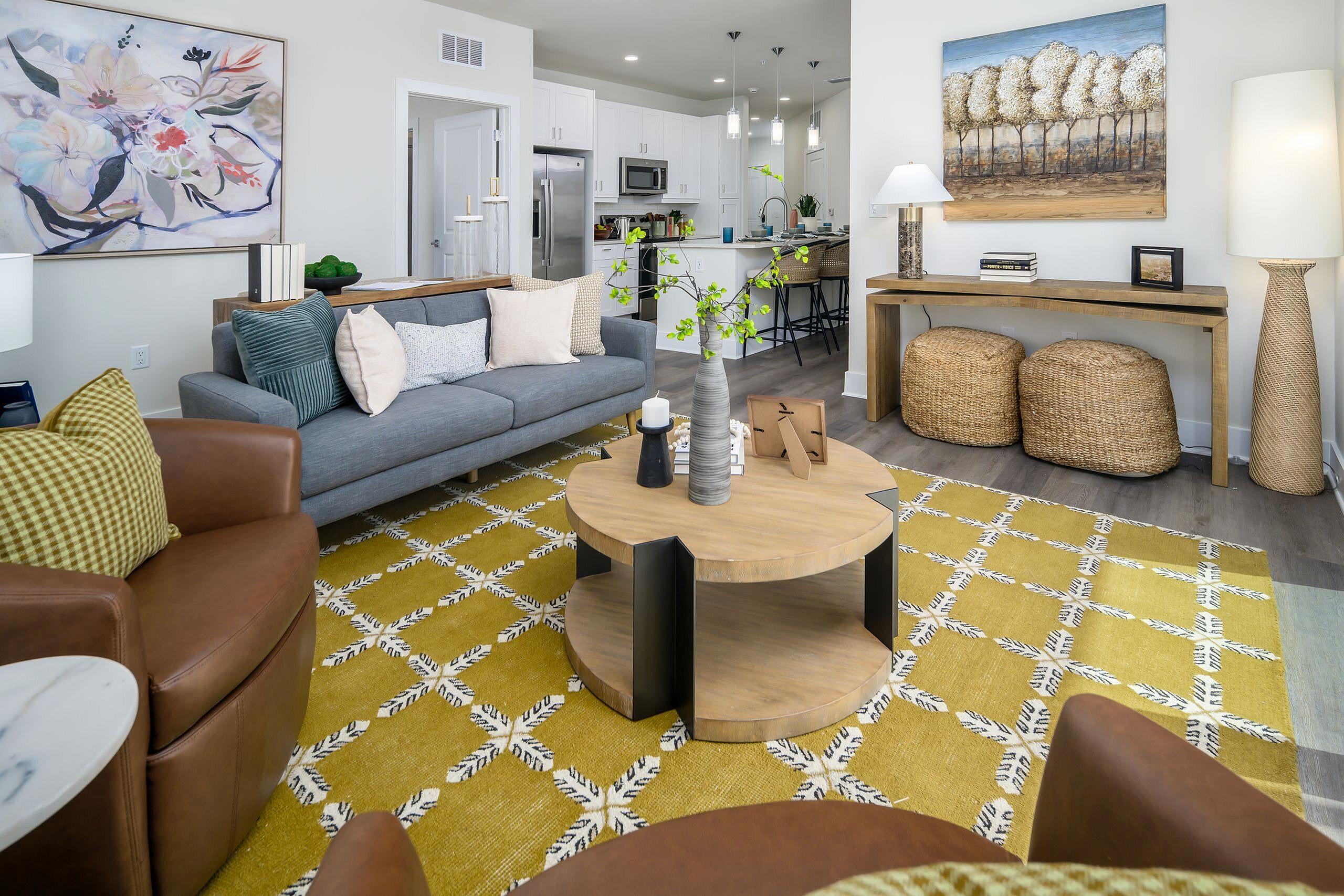 Inviting living room with modern decor, featuring a cozy gray sofa, stylish armchairs, and an elegant coffee table on a chic patterned rug, leading into a bright and open kitchen.